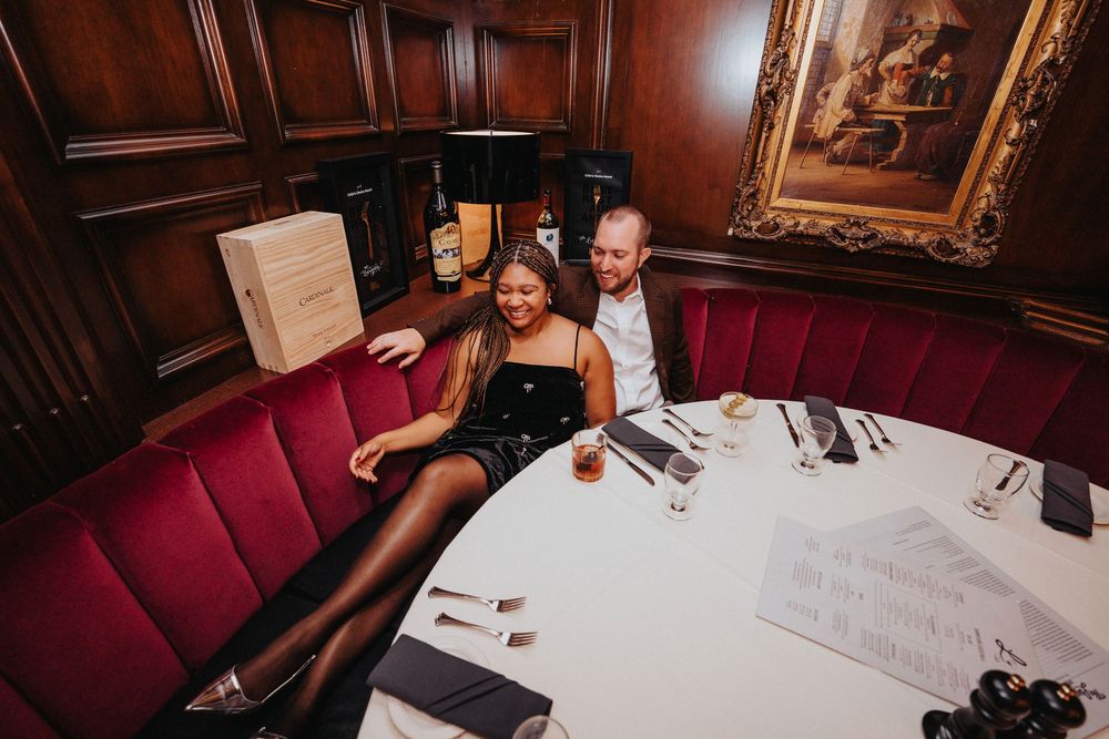 Elegant couple lounges at upscale restaurant booth with red velvet seating and wood-paneled walls.