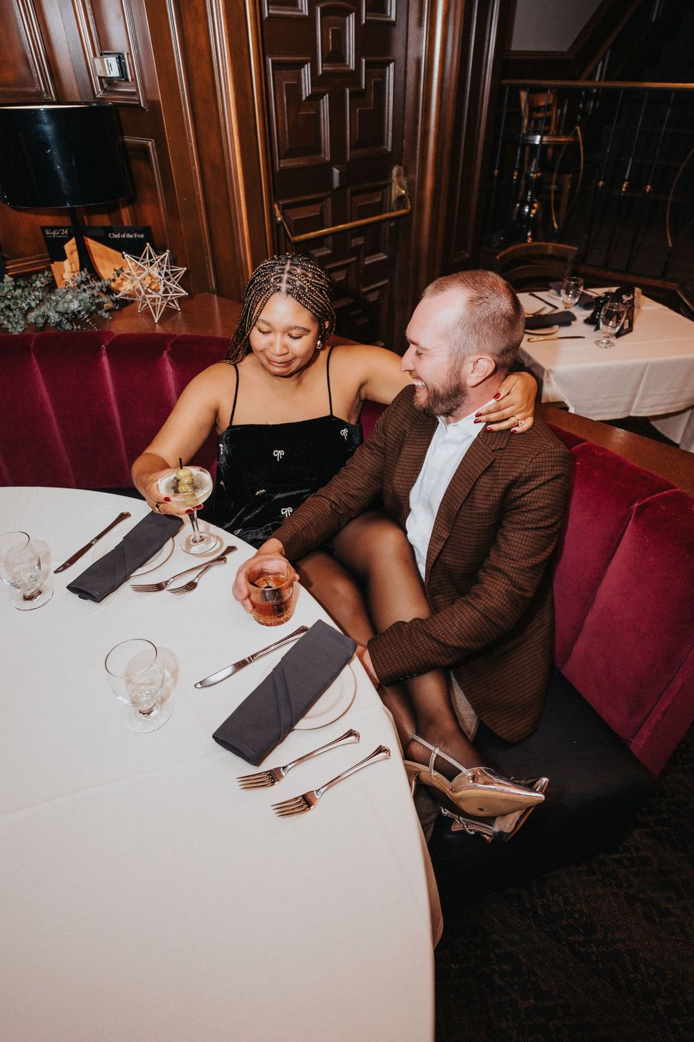 Couple enjoying drinks and conversation at an intimate restaurant table with red velvet seating.