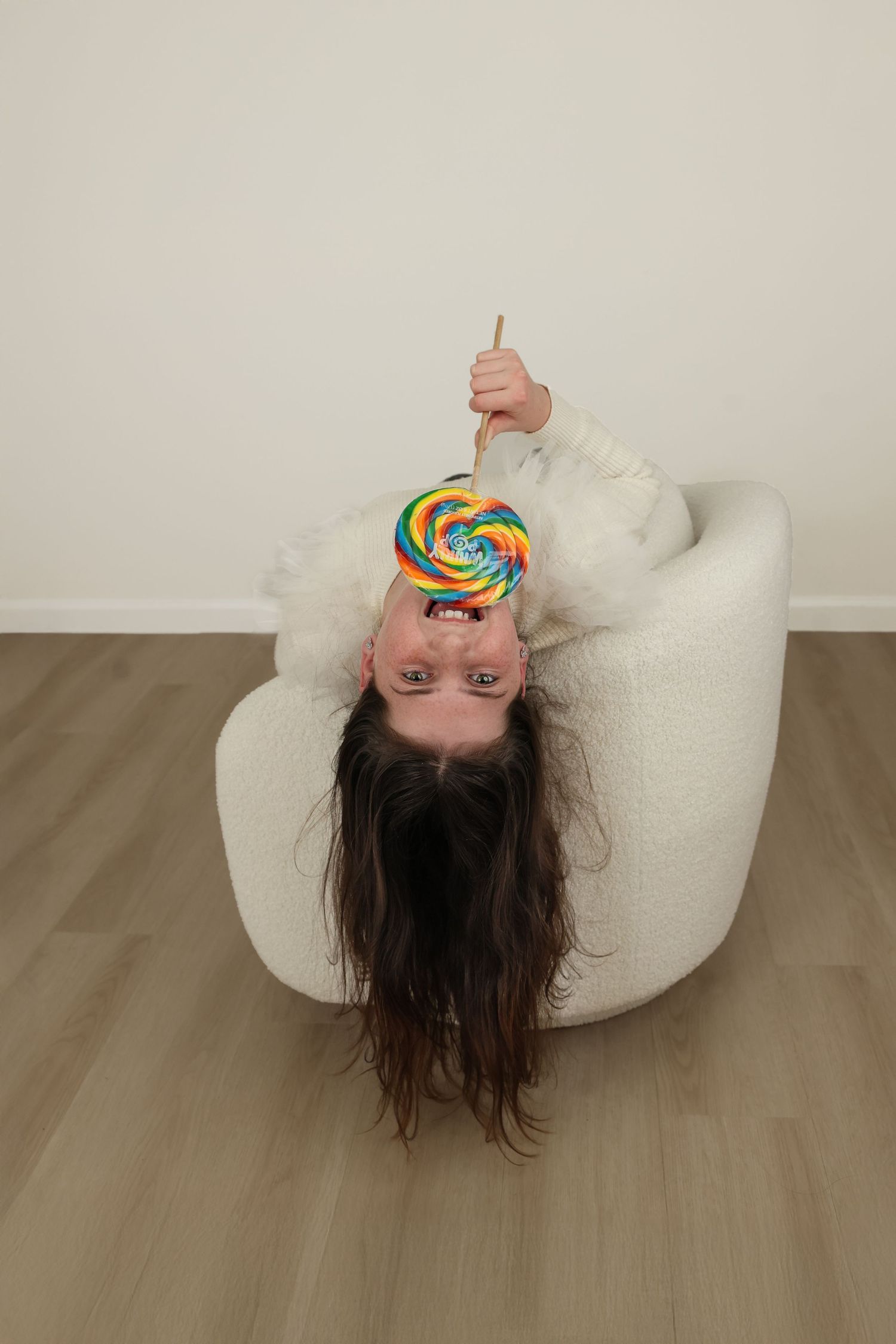 Playful studio portrait of a young girl with long brown hair hanging upside down on a white boucle chair holding a giant rainbow swirl lolly
