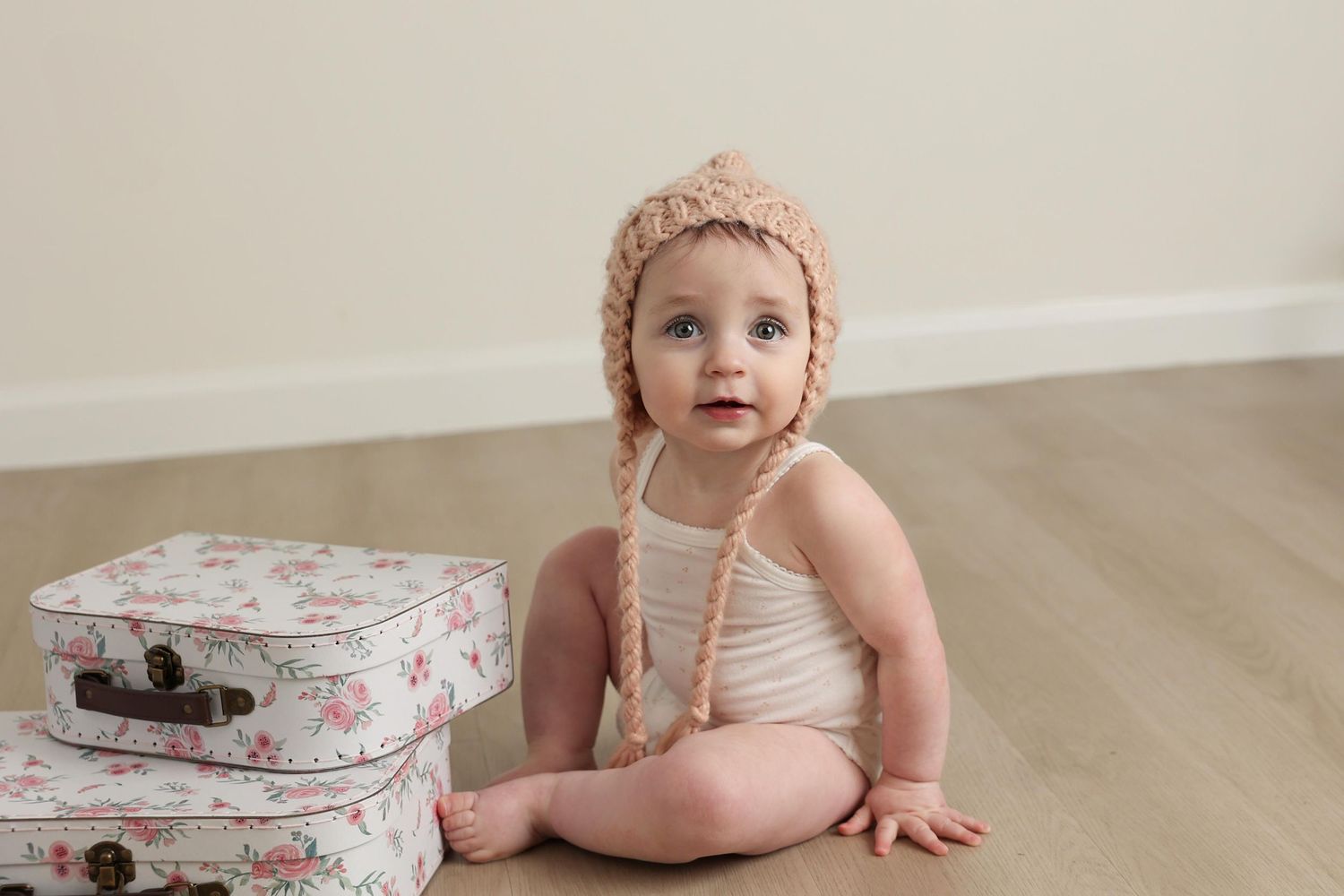 Studio sitter session portrait of a baby girl wearing a chunky knit bonnet sitting unassisted next to vintage floral suitcases