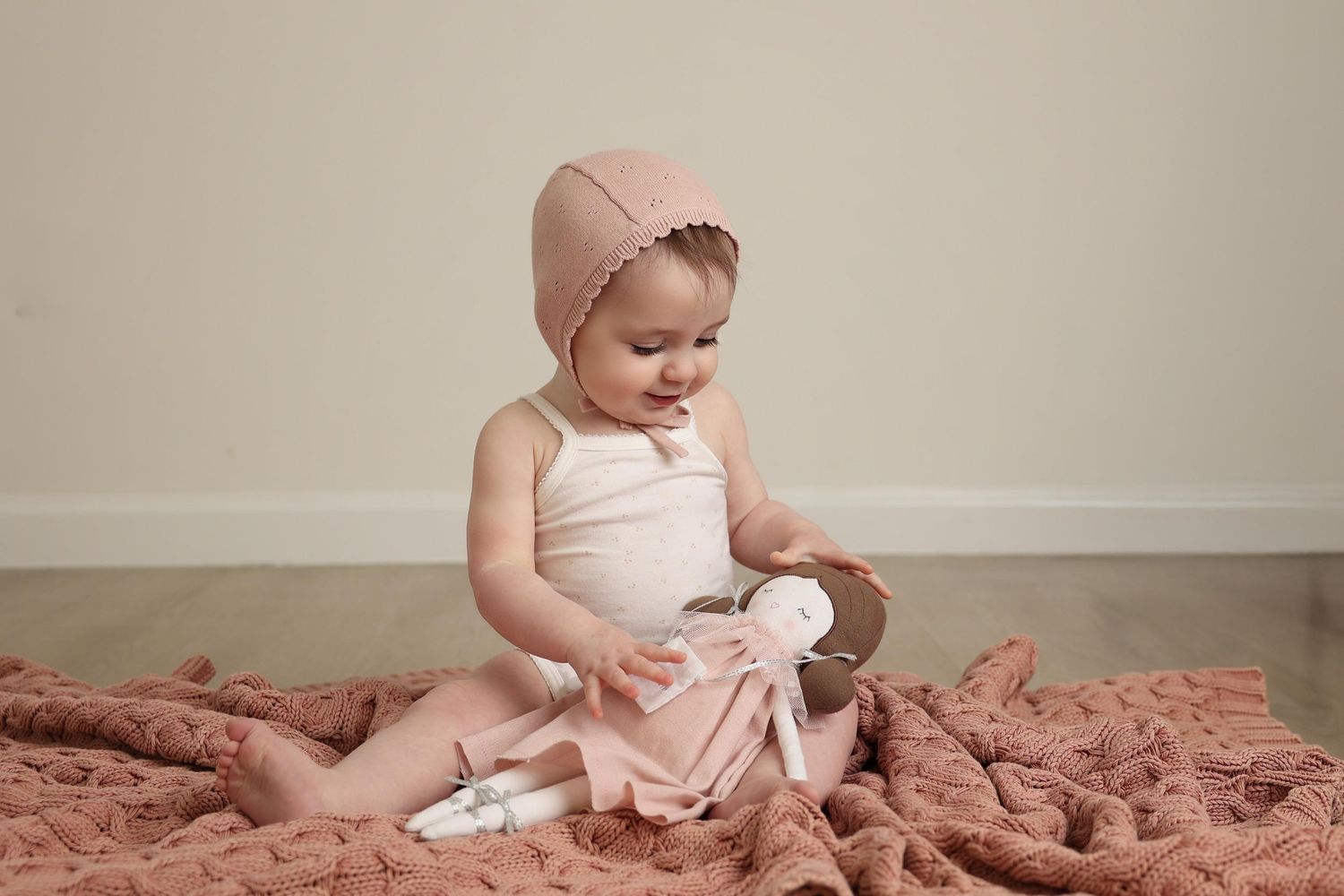 infant girl wearing a knot bonnet sitting on a soft pink blanket, playing with her pretty doll