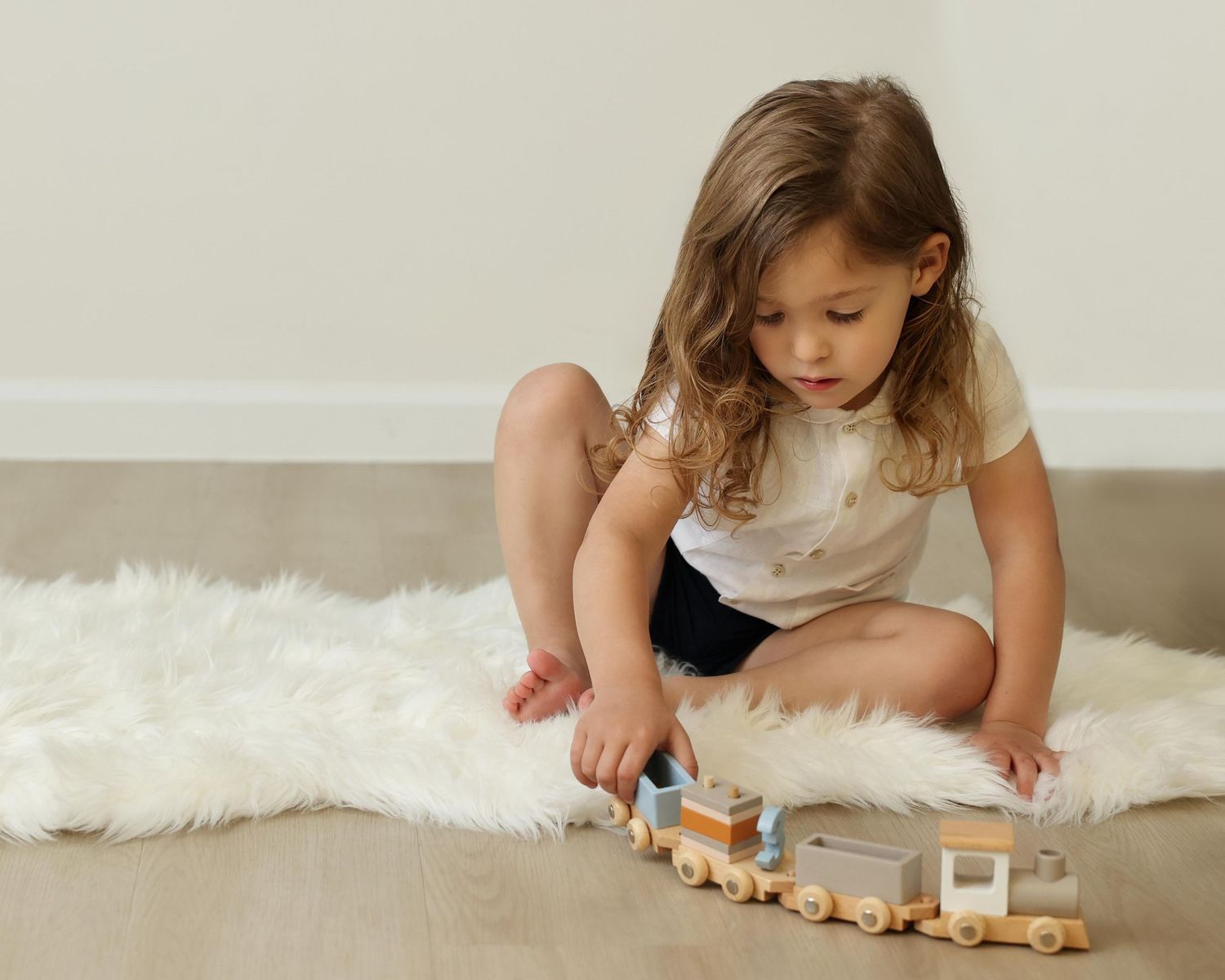 Candid photo of an upsherin boy sitting on a white rug pushing a toy train, highlighting his long curls