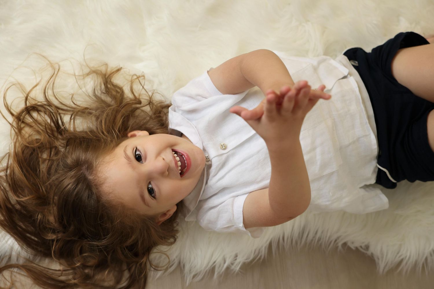 Overhead studio portrait of a happy upsherin boy lying on a white fur rug with his long curly hair fanned out around his head