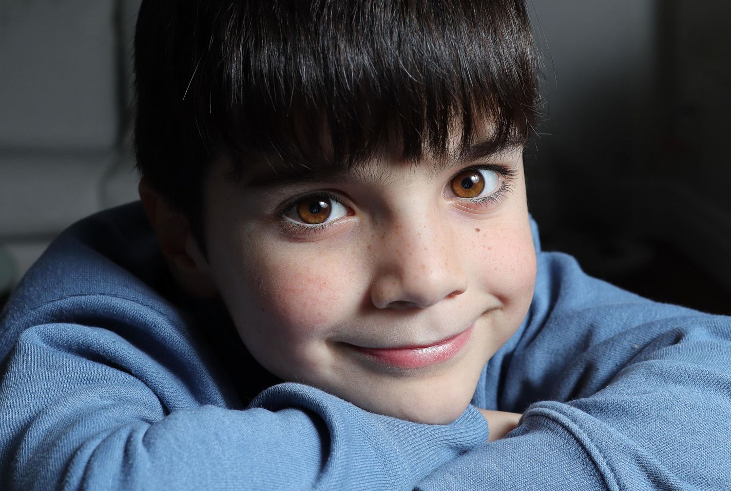 Close-up fine art studio portrait of a young boy with dark bangs and brown eyes smiling directly at the camera