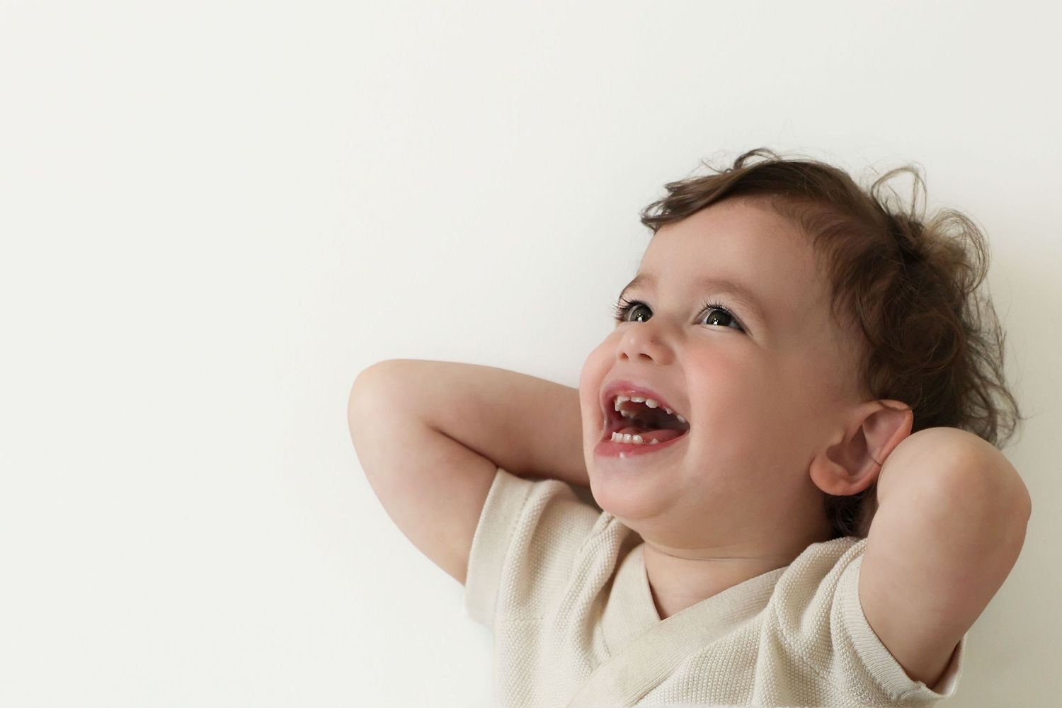 Candid personality shot of a toddler boy with curly hair laughing with his hands behind his head.