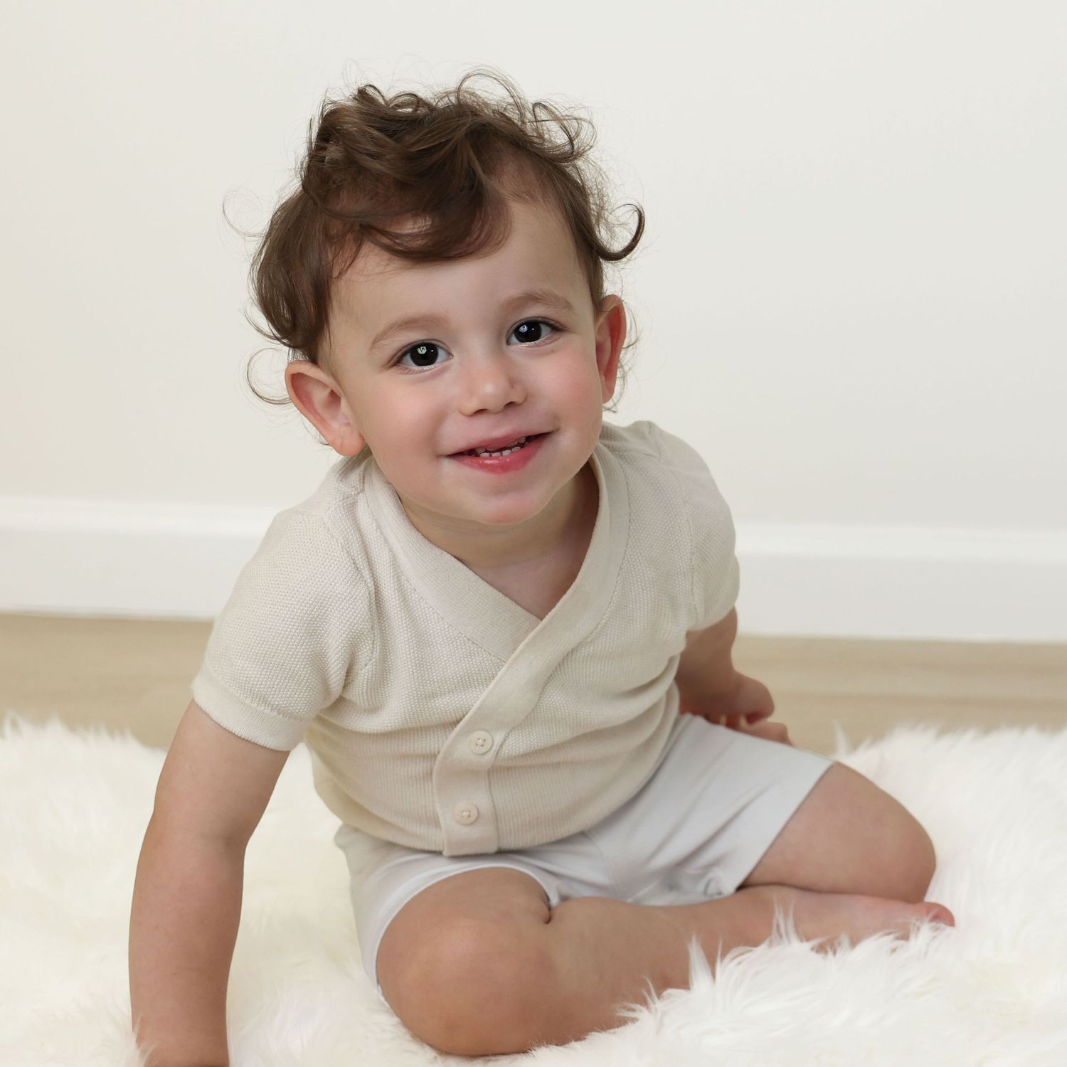 Studio milestone portrait of a smiling 1-year-old boy with dark curly hair sitting on a white fur rug