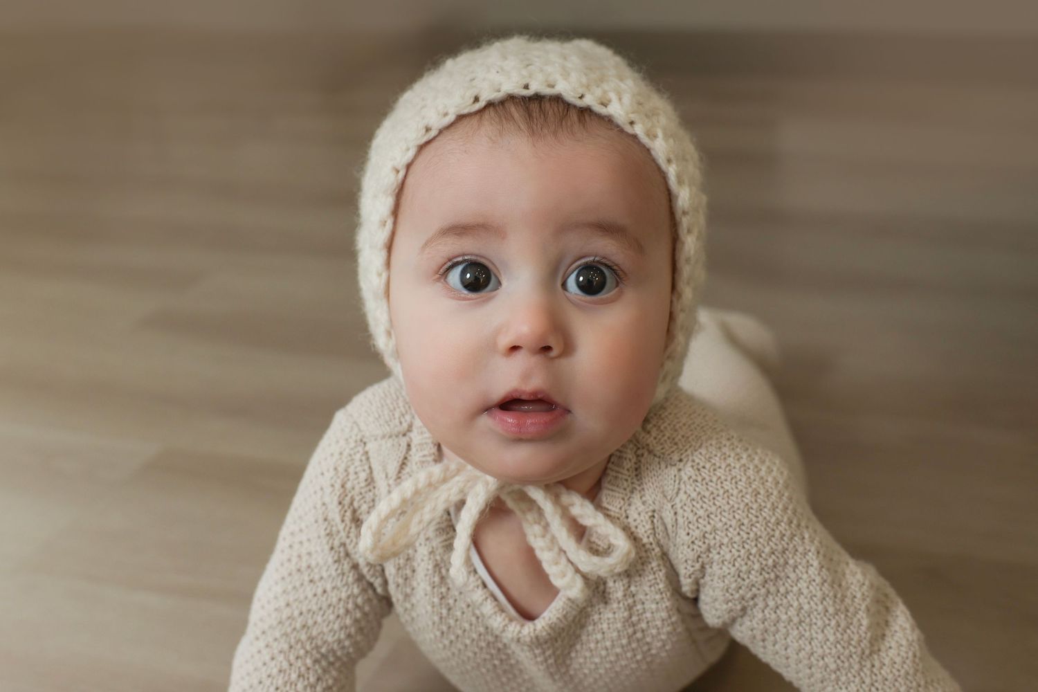 close up of a wide-eyed baby with a surprised expression wearing a knit hat and lying on a wood floor taken in Gitty Strauss Studio
