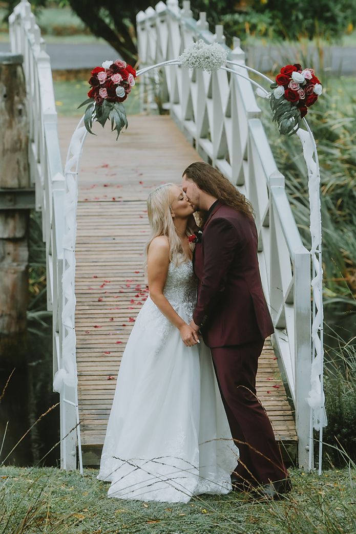 Romantic wedding couple shares intimate moments on a white wooden bridge decorated with red floral arrangements.