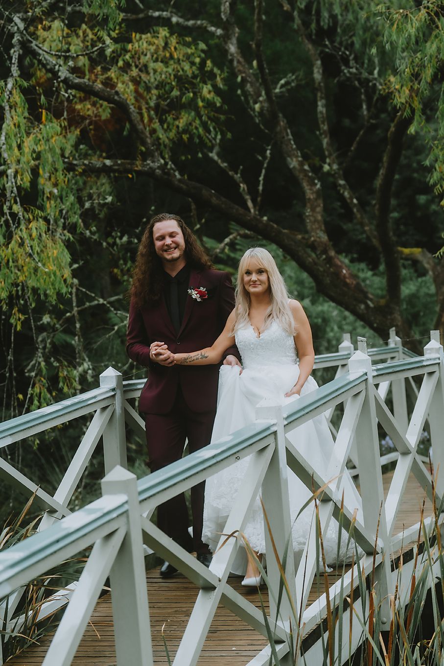 A couple walks together and shares a laugh while standing on a white wooden bridge in a natural setting.