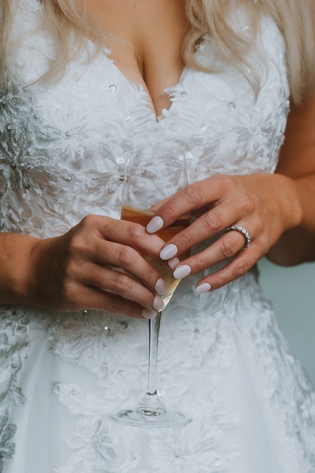 Close-up of bride's hands with white manicure and wedding ring holding champagne glass against lace wedding dress.