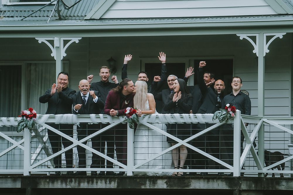 A large group of people stand on a white porch railing in formal attire at an outdoor event.