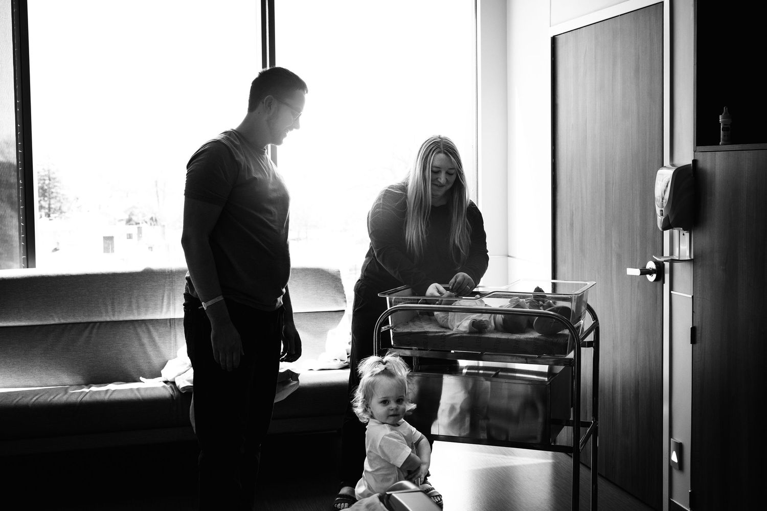 A toddler explores while adults attend to tasks in a bright hospital room.