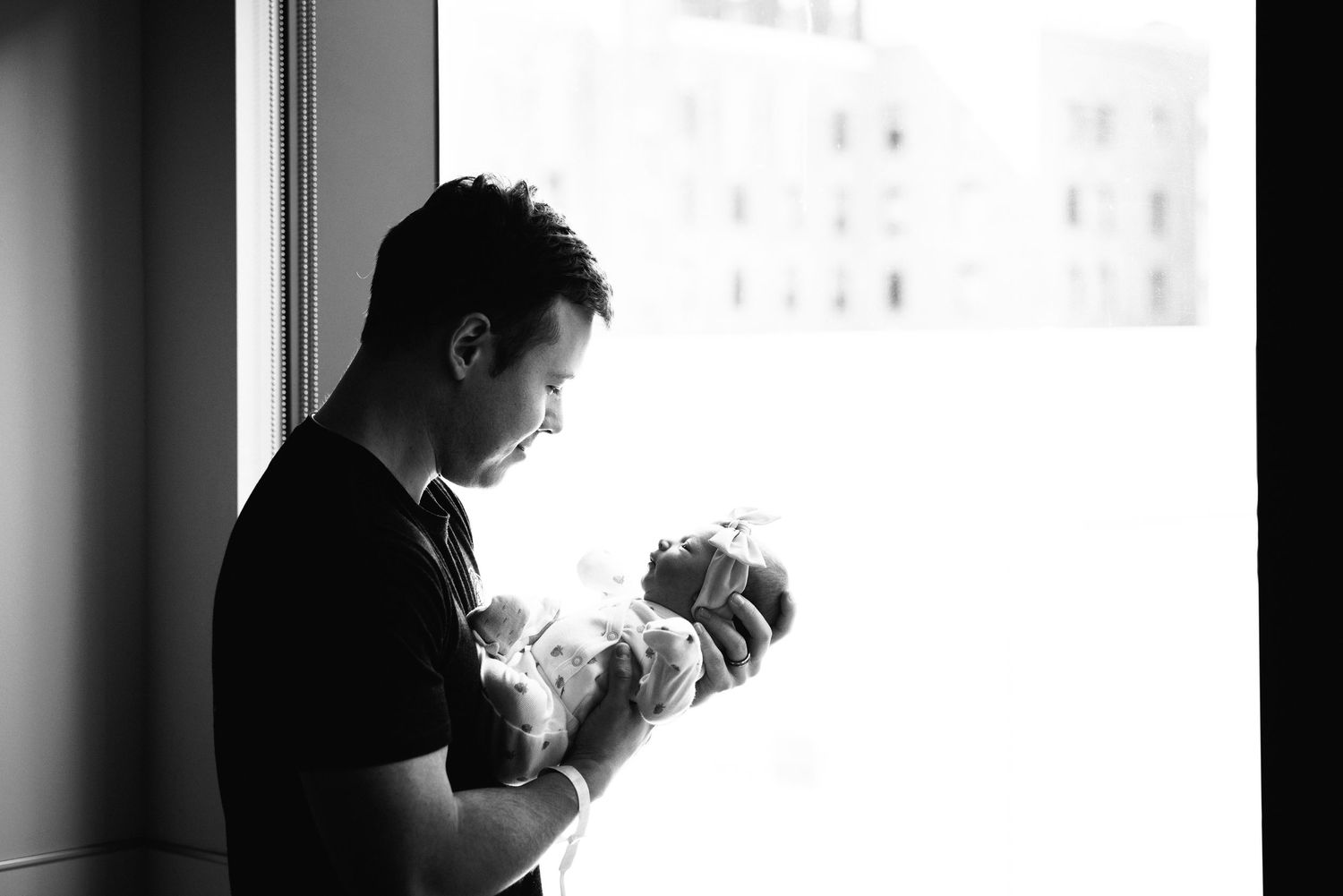 A silhouetted figure stands by a window in black and white, tenderly holding a newborn baby.