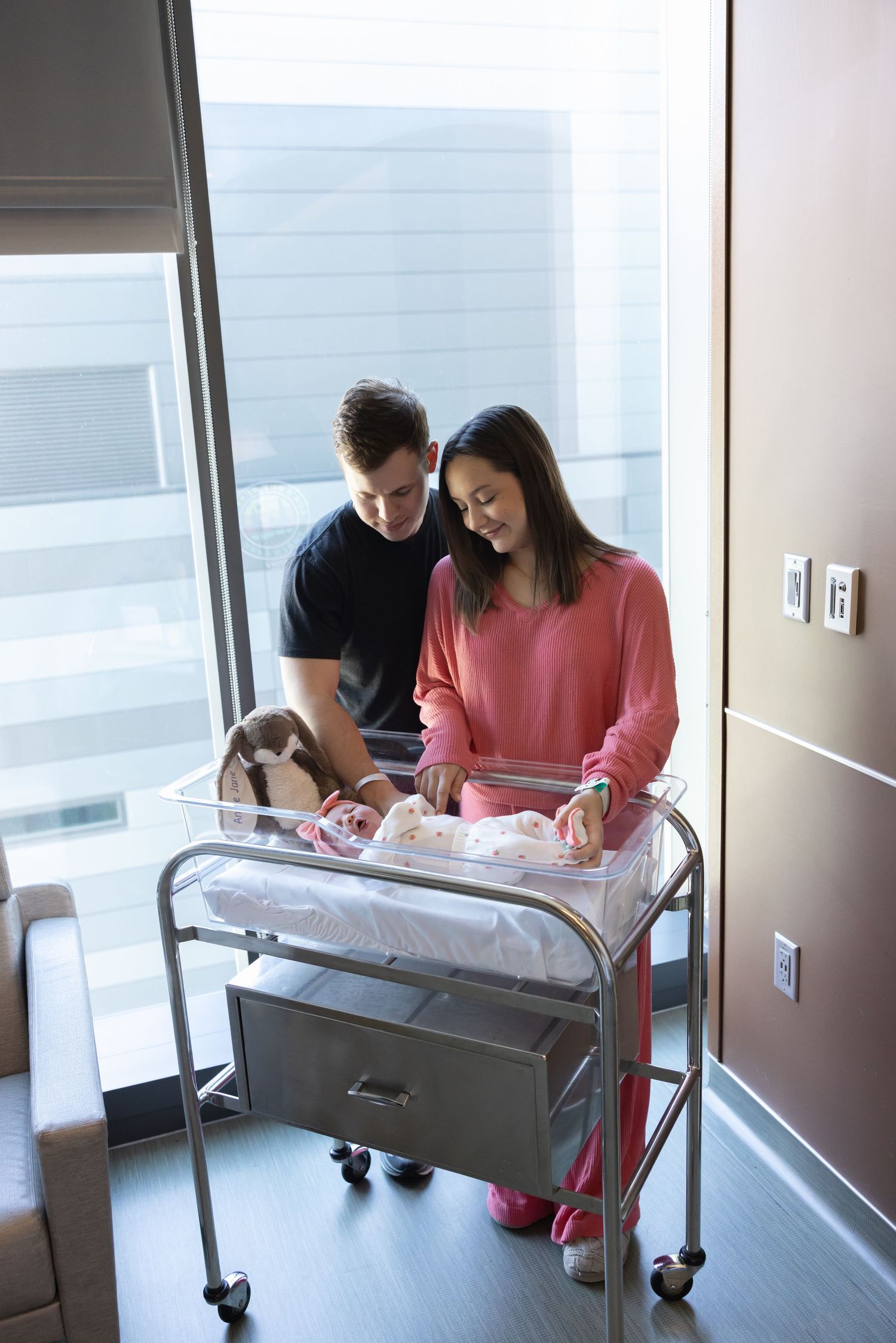 New parents stand together looking at their newborn baby resting in a hospital bassinet.