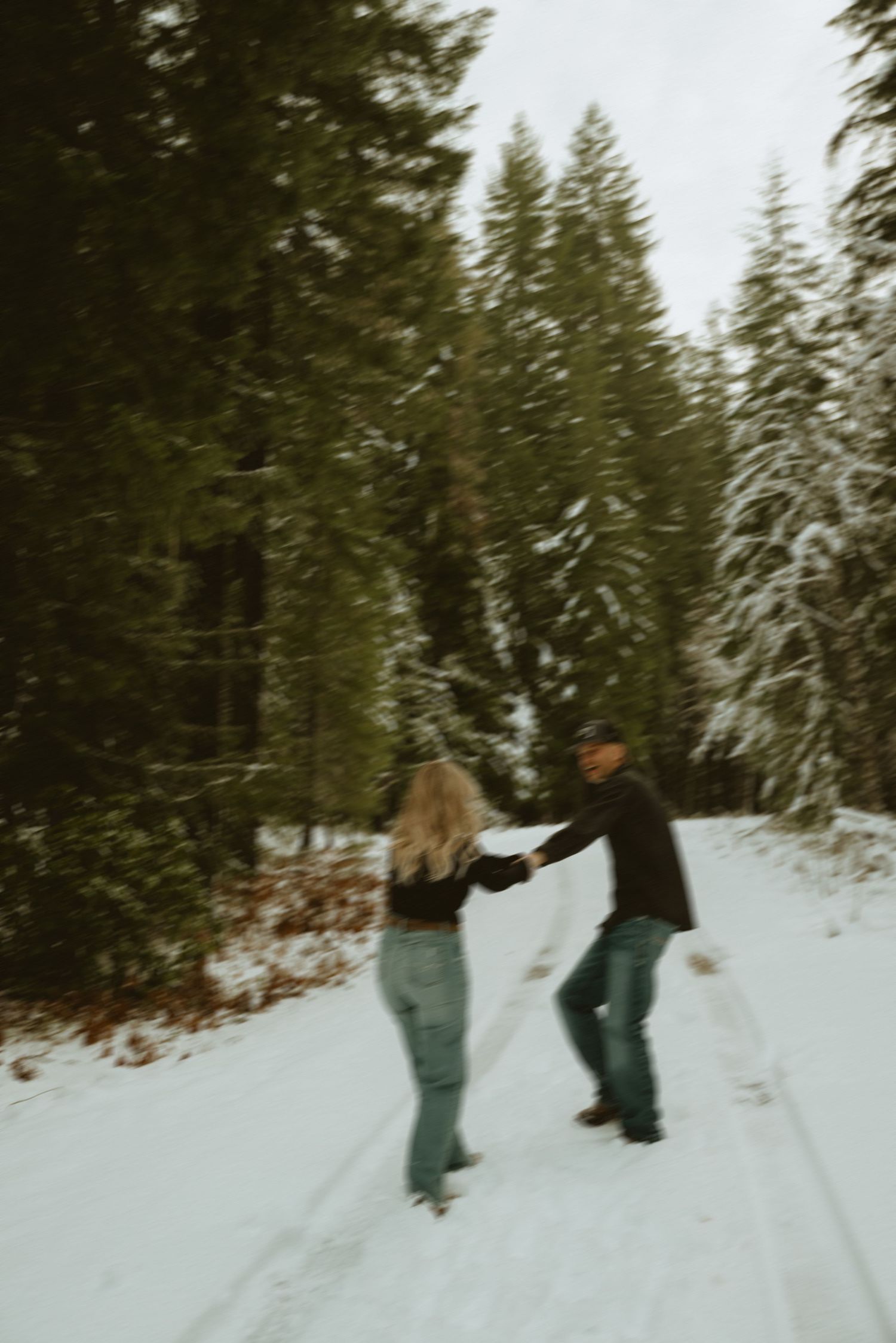 A couple embraces in a snowy forest setting with tall evergreen trees in the background.