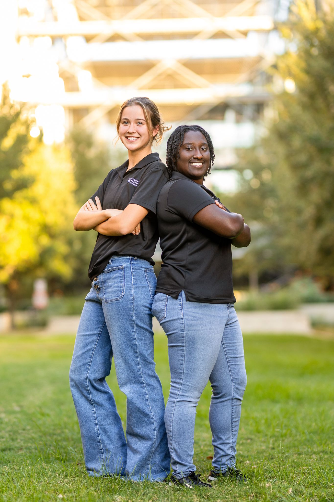 Two friends in matching outfits sitting back to back on grass during a sunny day outdoors.