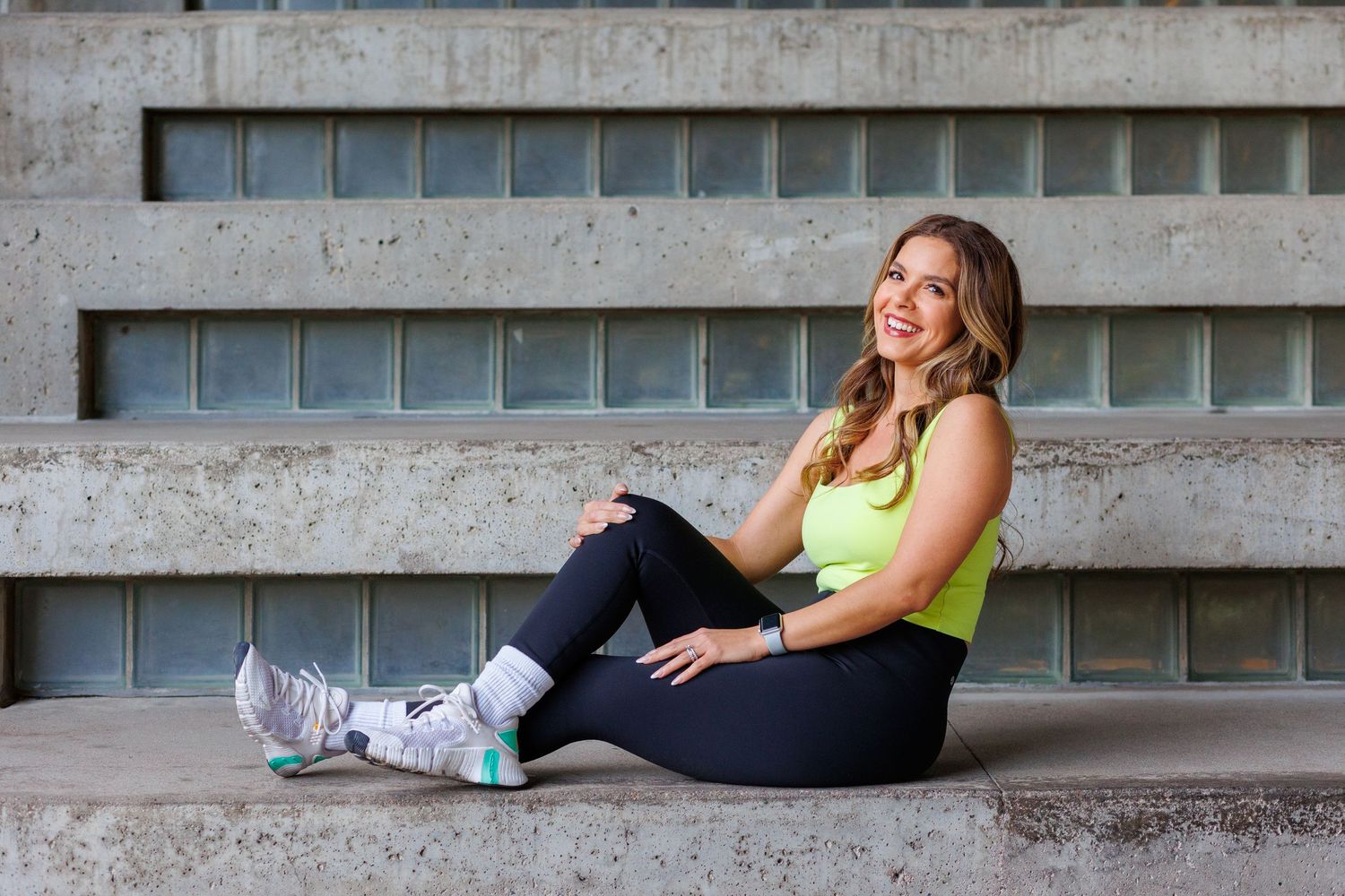 Smiling woman in yellow top and black leggings sits on concrete steps in athletic wear.