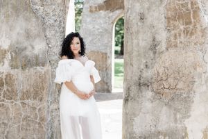 A person in an elegant white off-shoulder dress poses gracefully against weathered stone walls with arched windows.