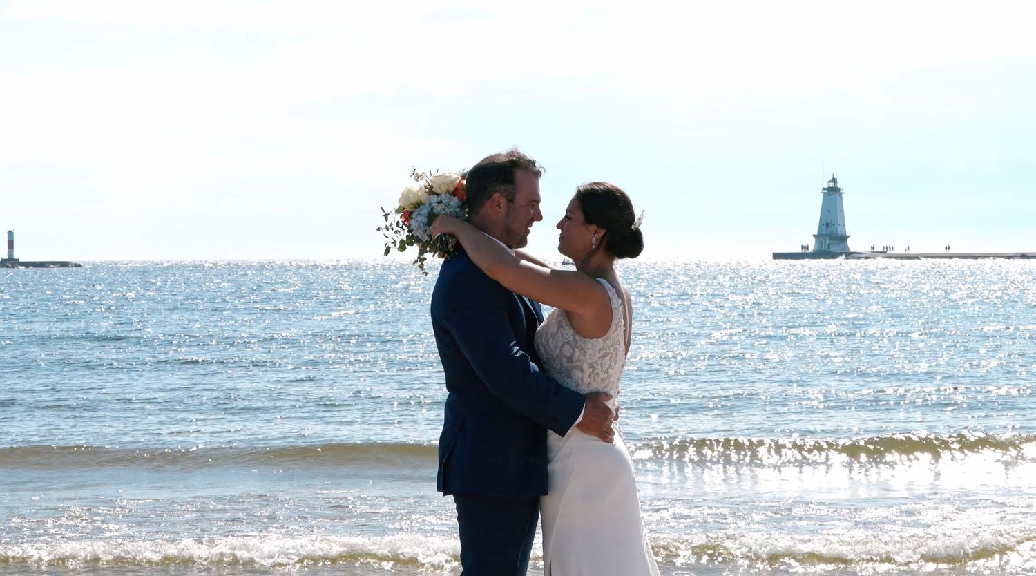 A couple shares a romantic moment on the beach with a lighthouse visible in the distance on a sunny day.