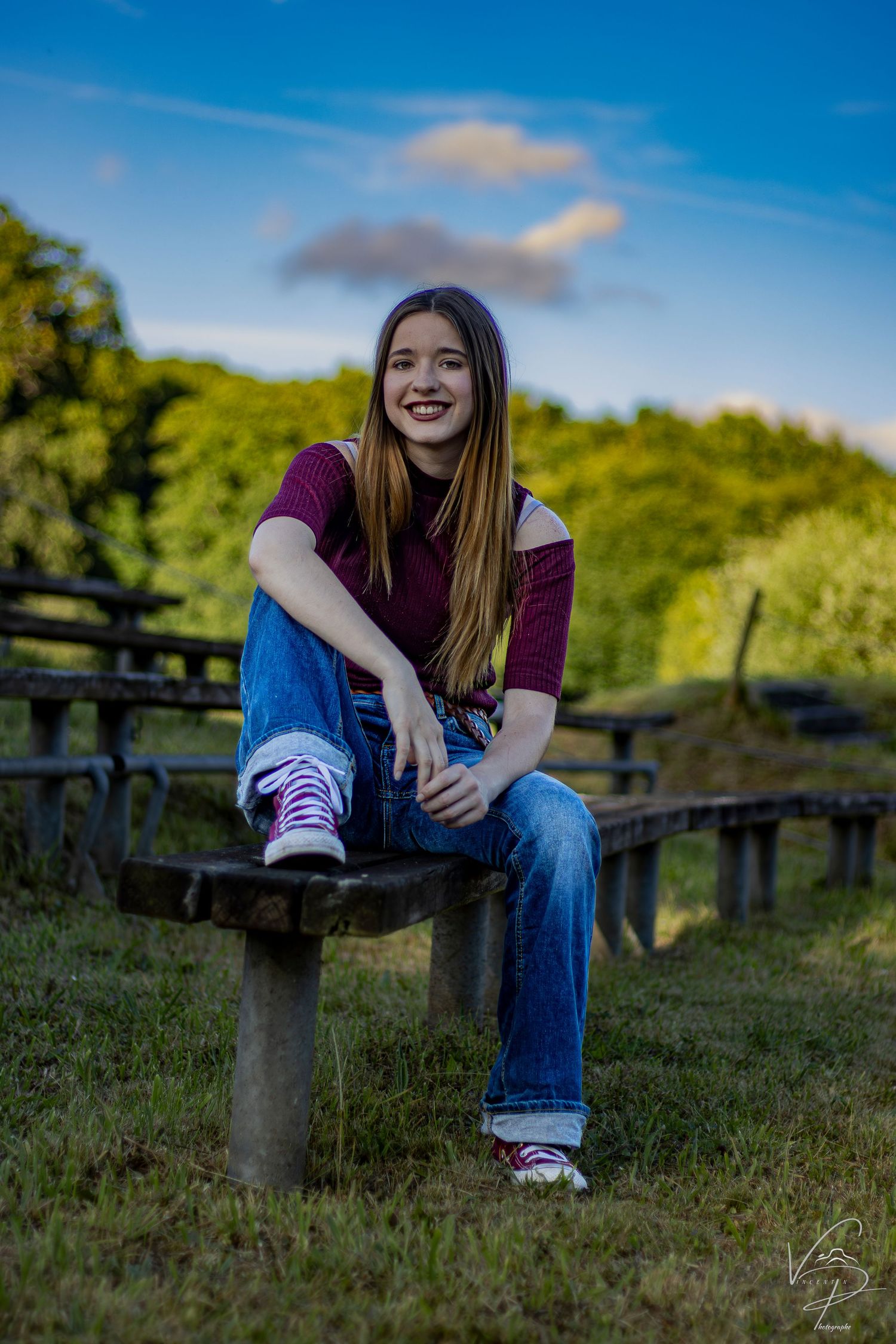Someone in a burgundy top and jeans sits casually on a wooden bench outdoors on a sunny day with green foliage behind.