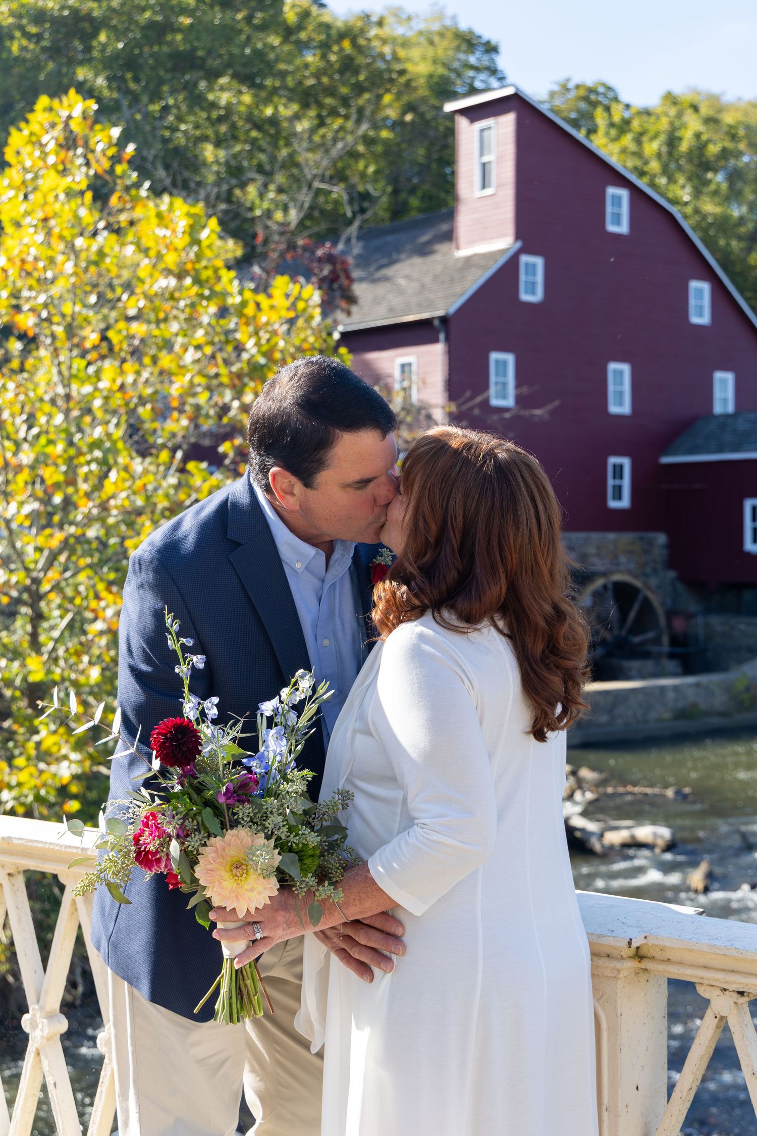 A romantic moment captured in front of a rustic red mill building with a waterwheel and autumn foliage.