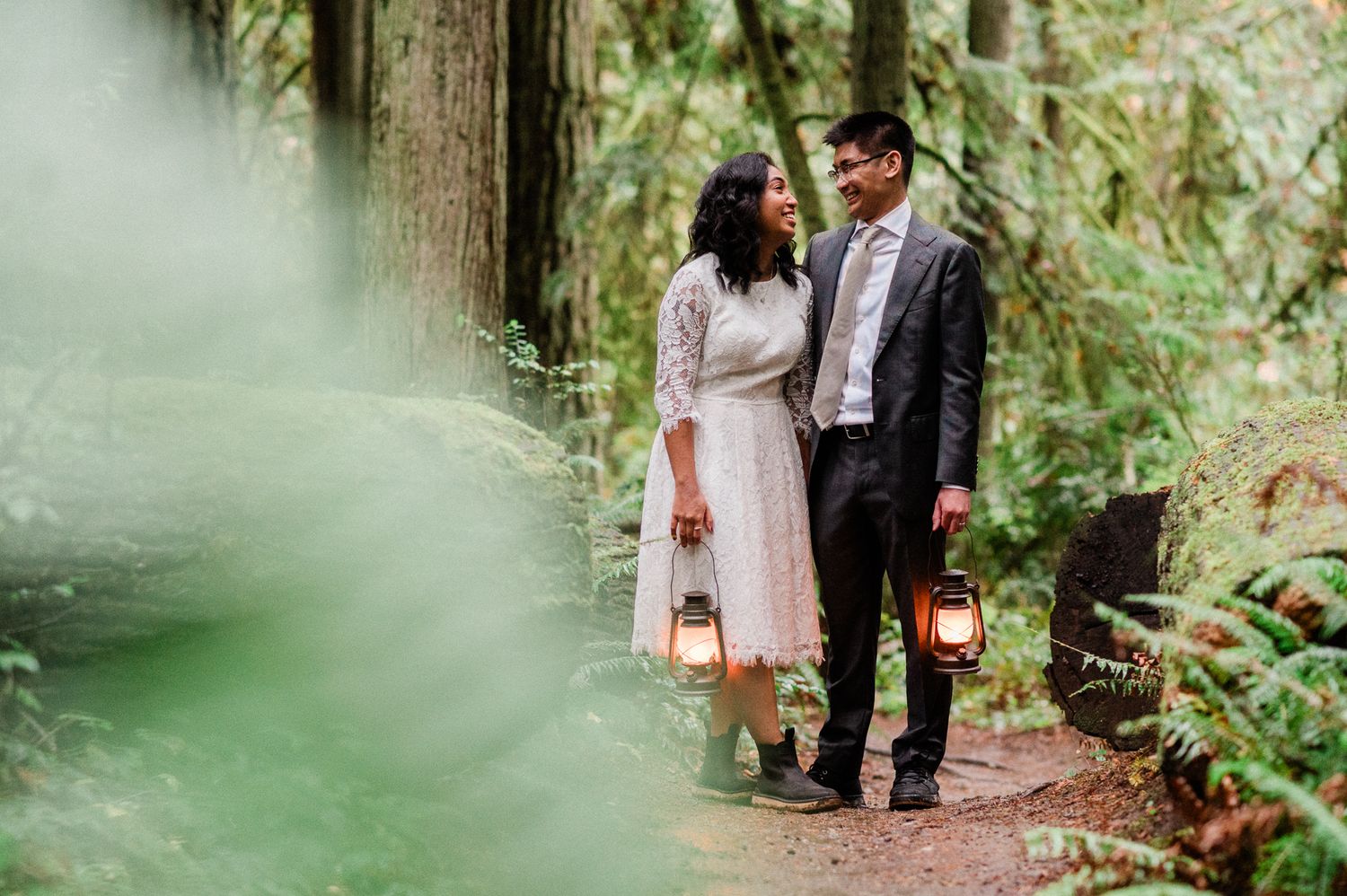 A couple in wedding attire walks through a lush forest path surrounded by green foliage on Bainbridge Island