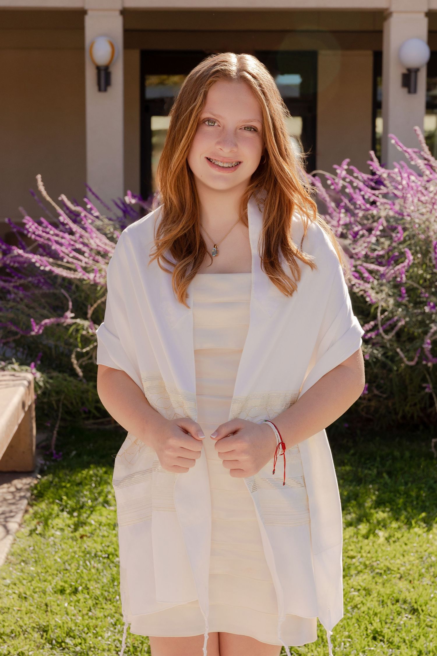 A person in a white outfit poses outdoors against a backdrop of purple flowering plants and columns.