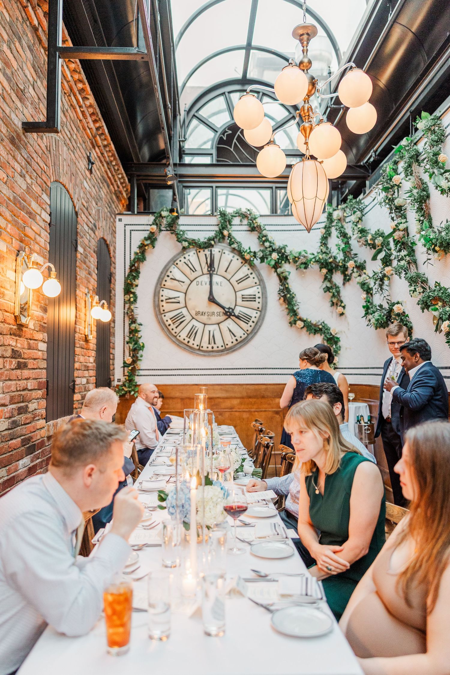 Elegant wedding reception dinner with guests seated at a long table under vintage clock and hanging lights with greenery decor.