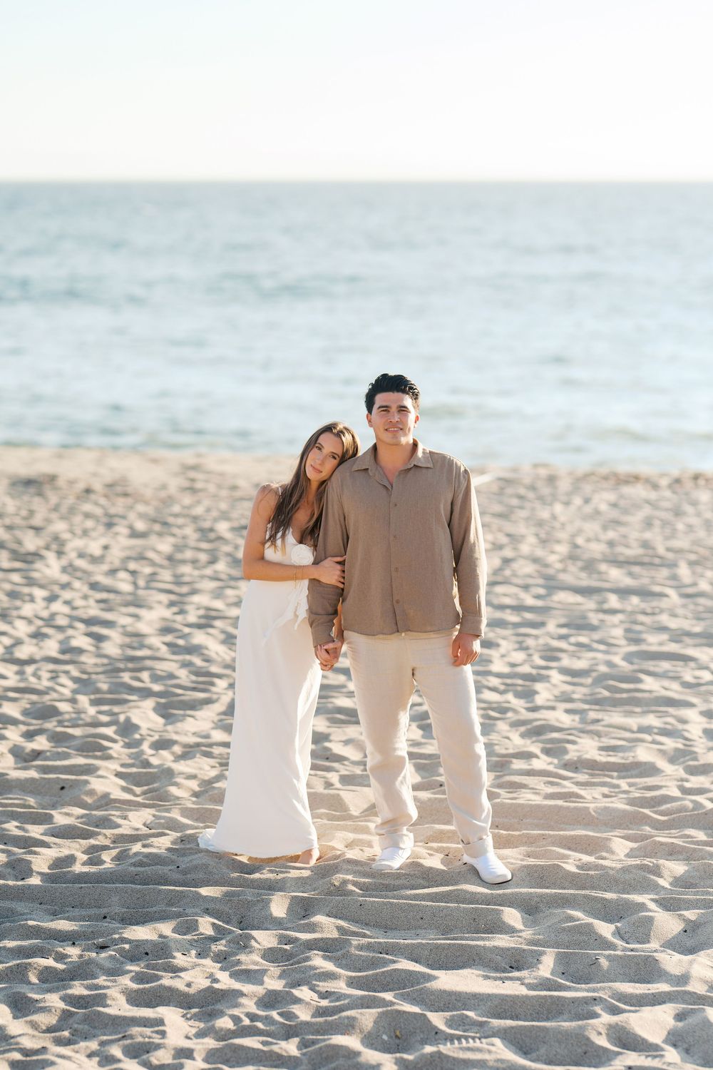 A couple in coordinated white and beige outfits shares romantic moments together on a sunny beach.