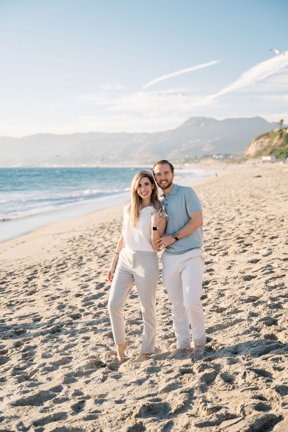 A couple wearing coordinated white outfits plays with their dog on a sunny beach at sunset.