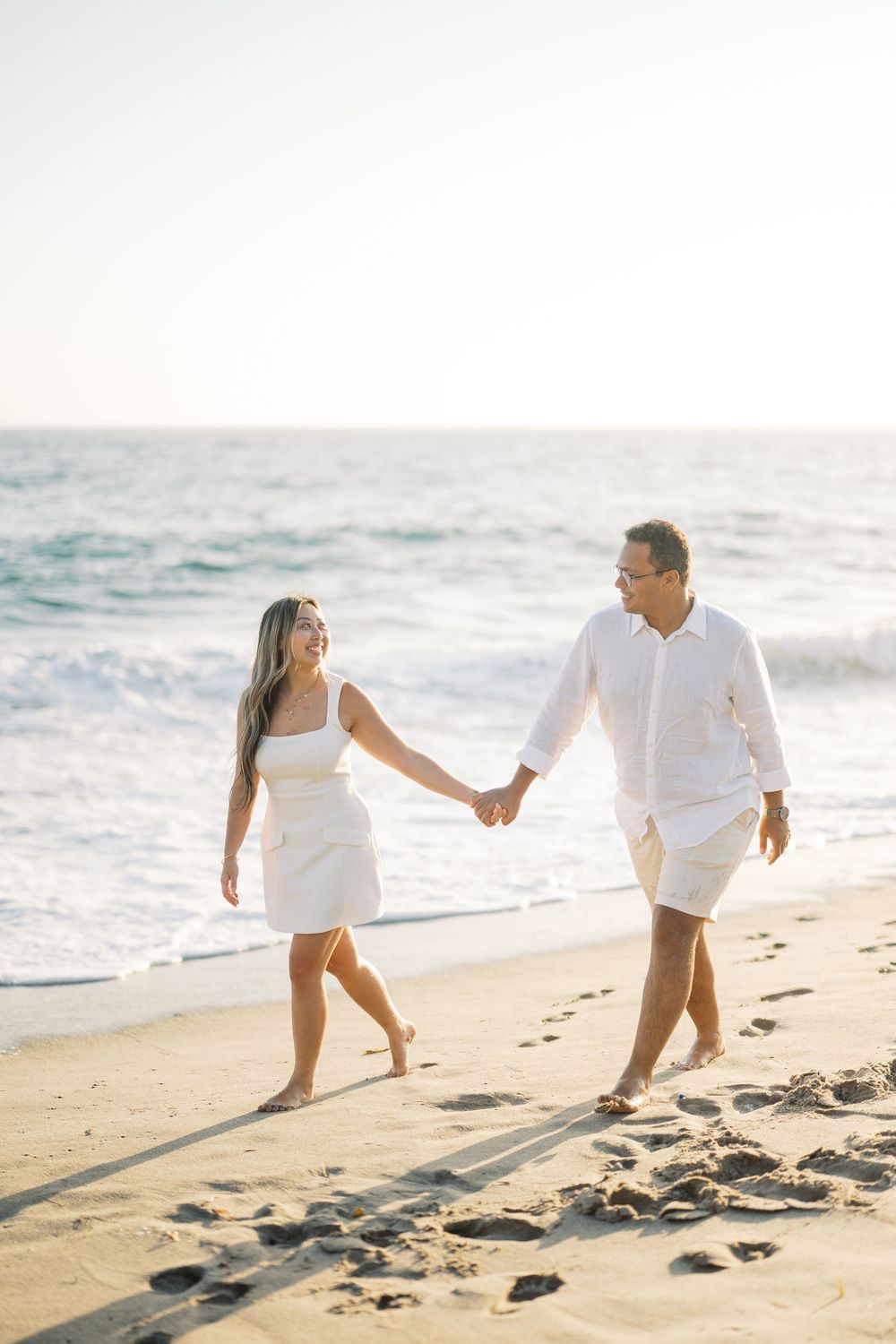 A couple walks hand in hand along a sunny beach shoreline during golden hour, creating romantic silhouettes in the sand.
