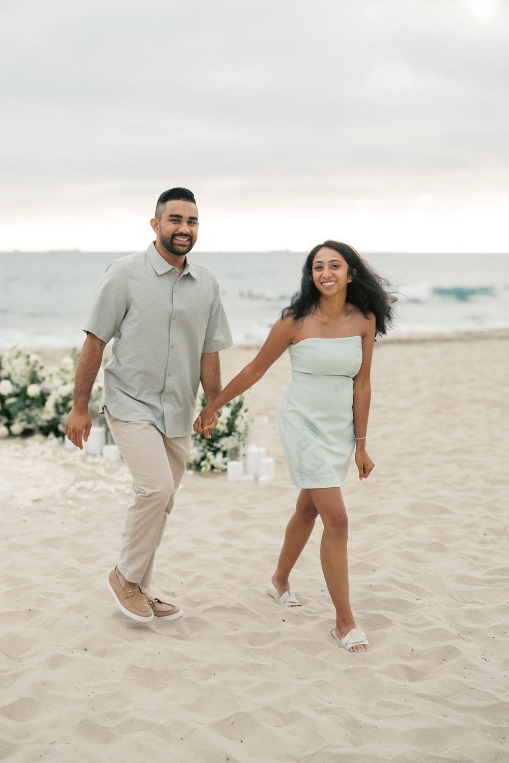 A couple in light colored outfits walking playfully on a sandy beach with ocean waves in the background.
