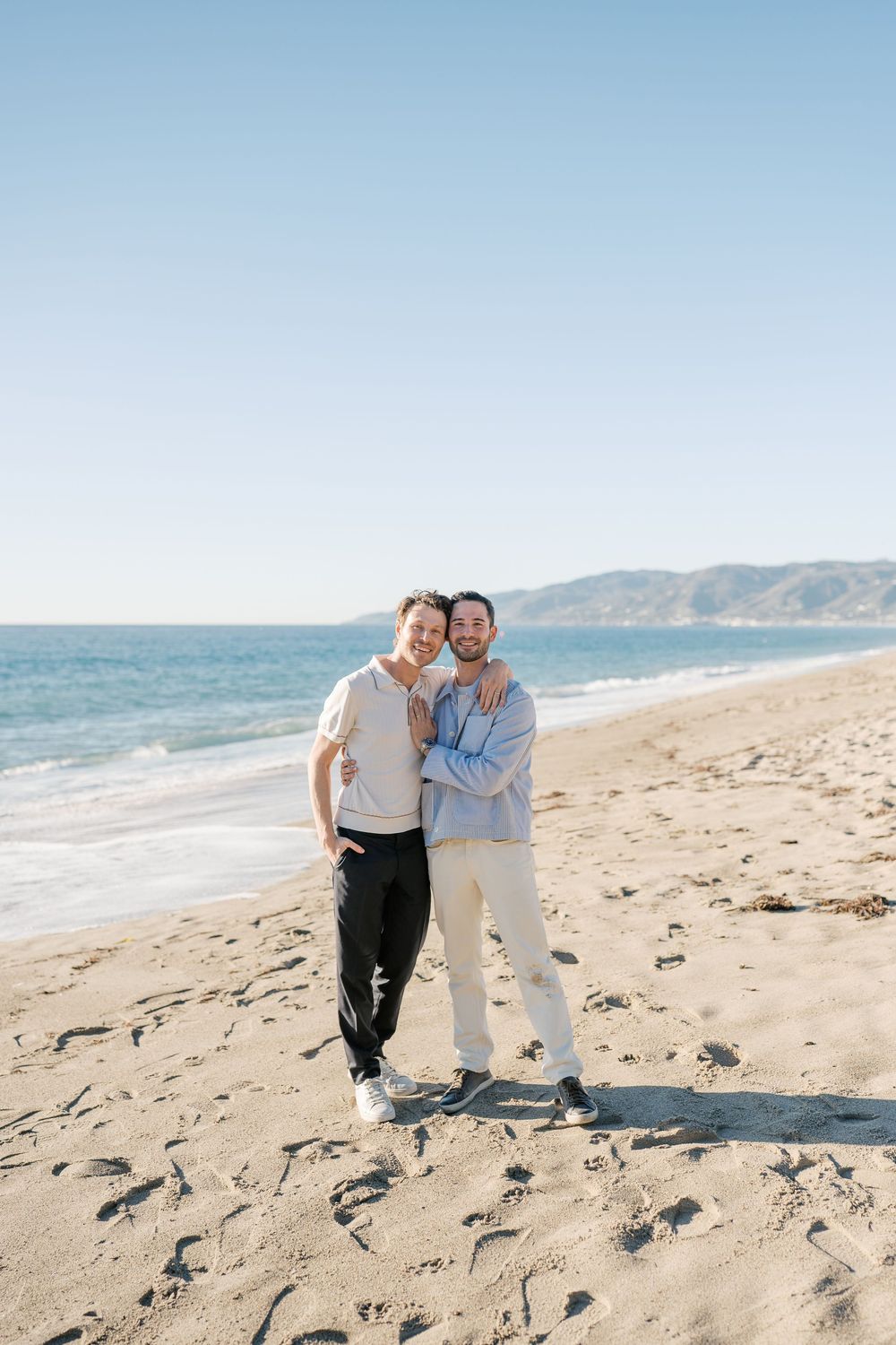 A couple embraces while standing on a sunny beach with mountains and ocean waves in the background.