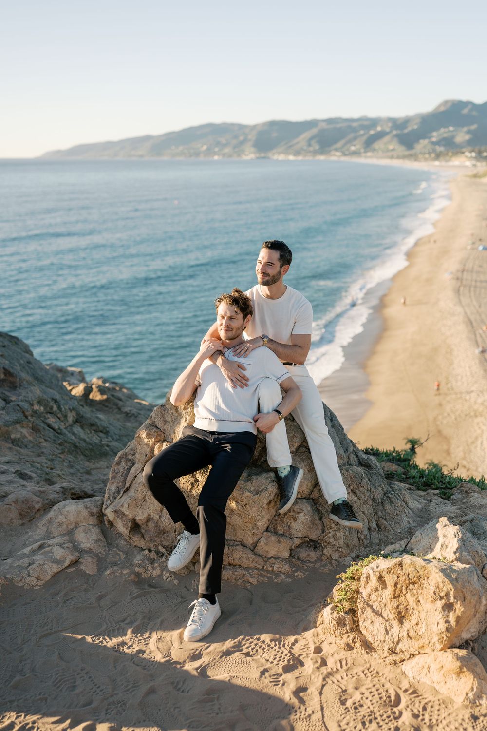 Couple enjoying a romantic moment on coastal cliffs overlooking a scenic beach and ocean view at sunset.