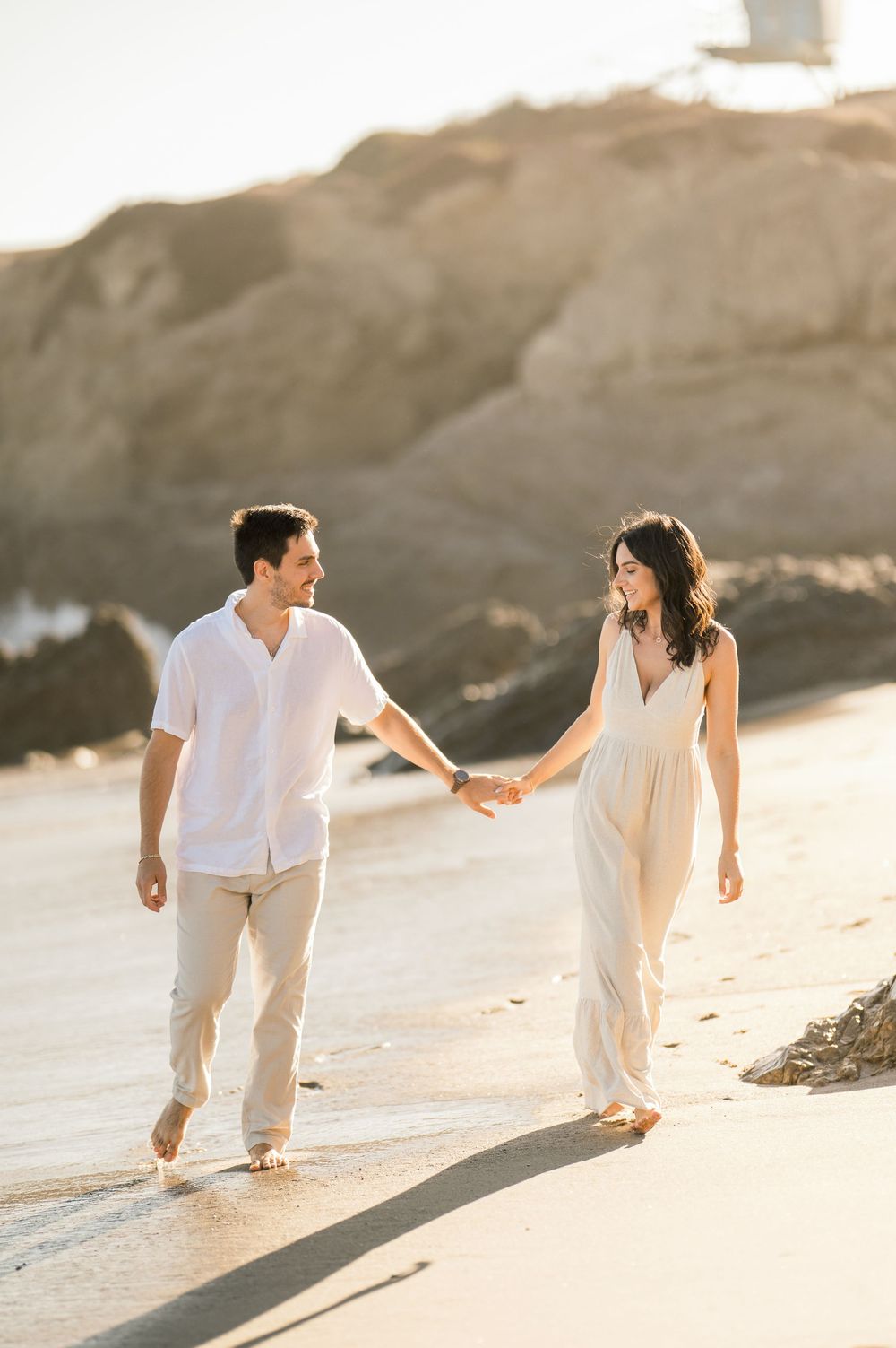 A couple in white outfits walks hand in hand along a sandy beach at sunset, creating a romantic silhouette.