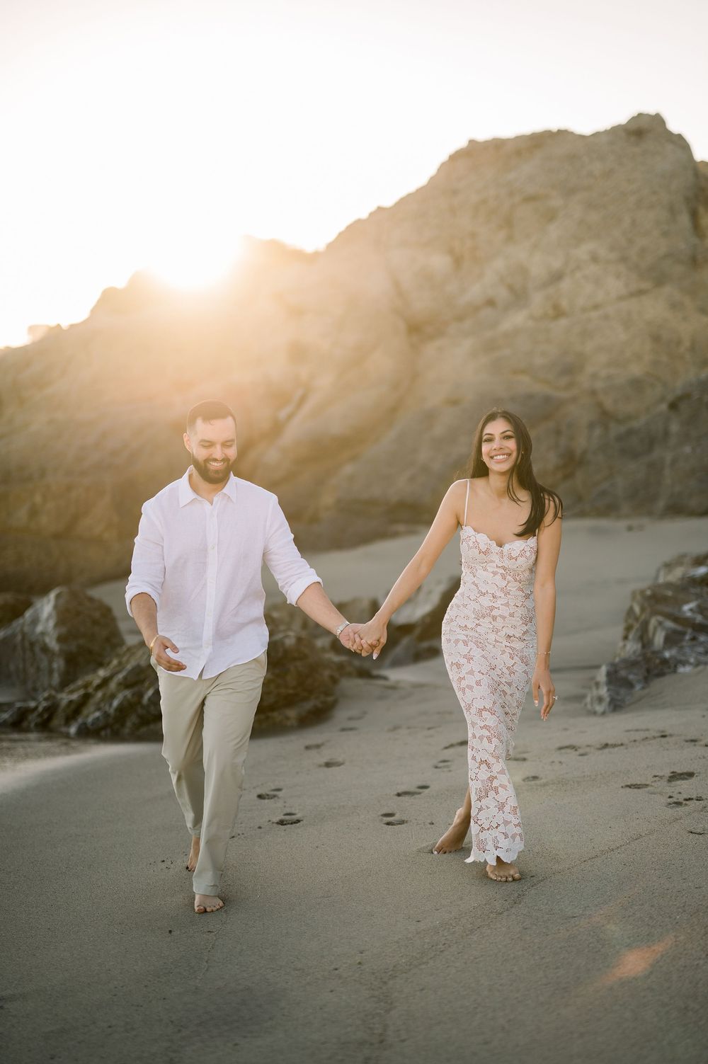 A couple walks hand in hand on a beach at sunset with rocky cliffs in the background.
