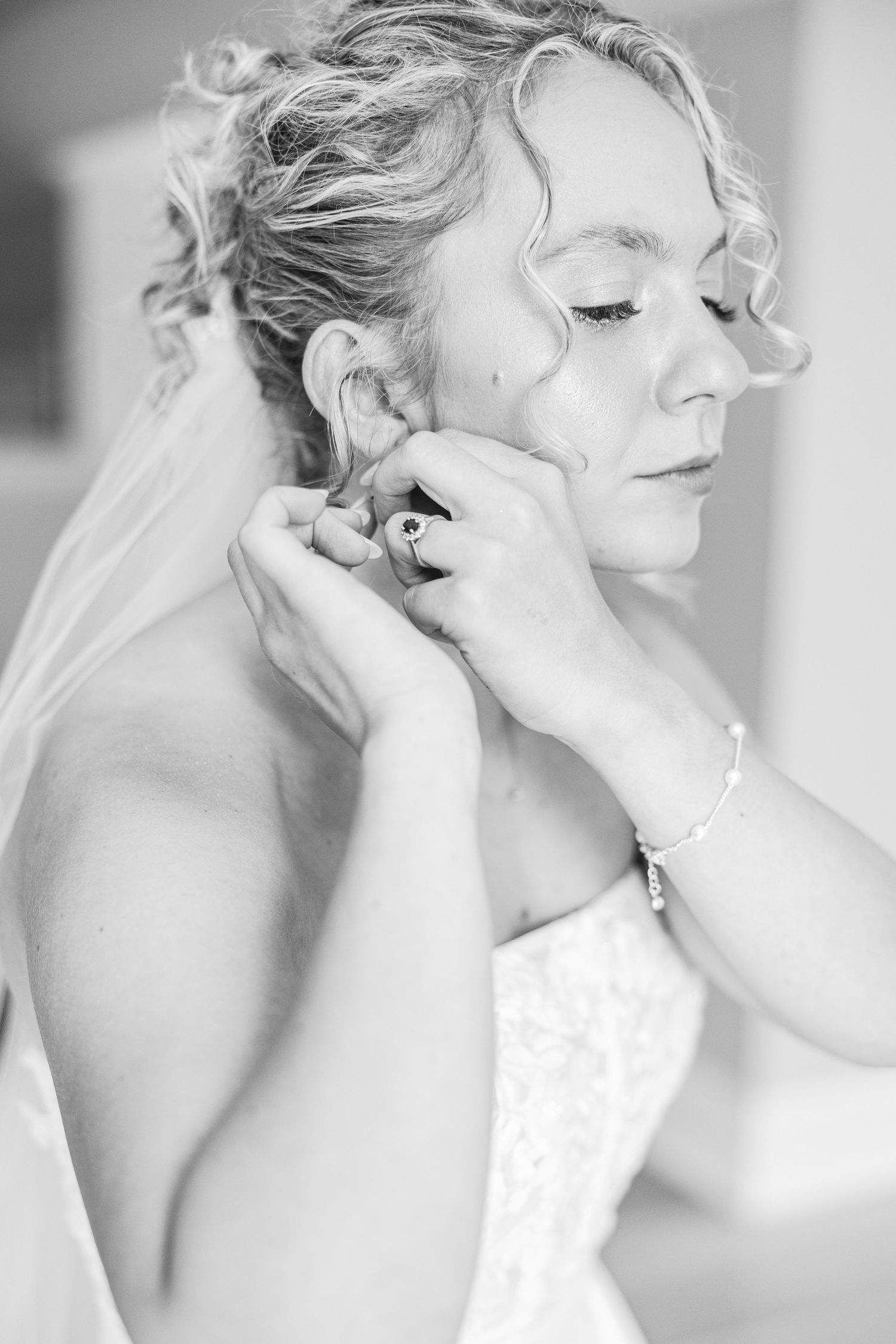 Black and white close-up of someone putting on earrings while wearing a wedding veil.