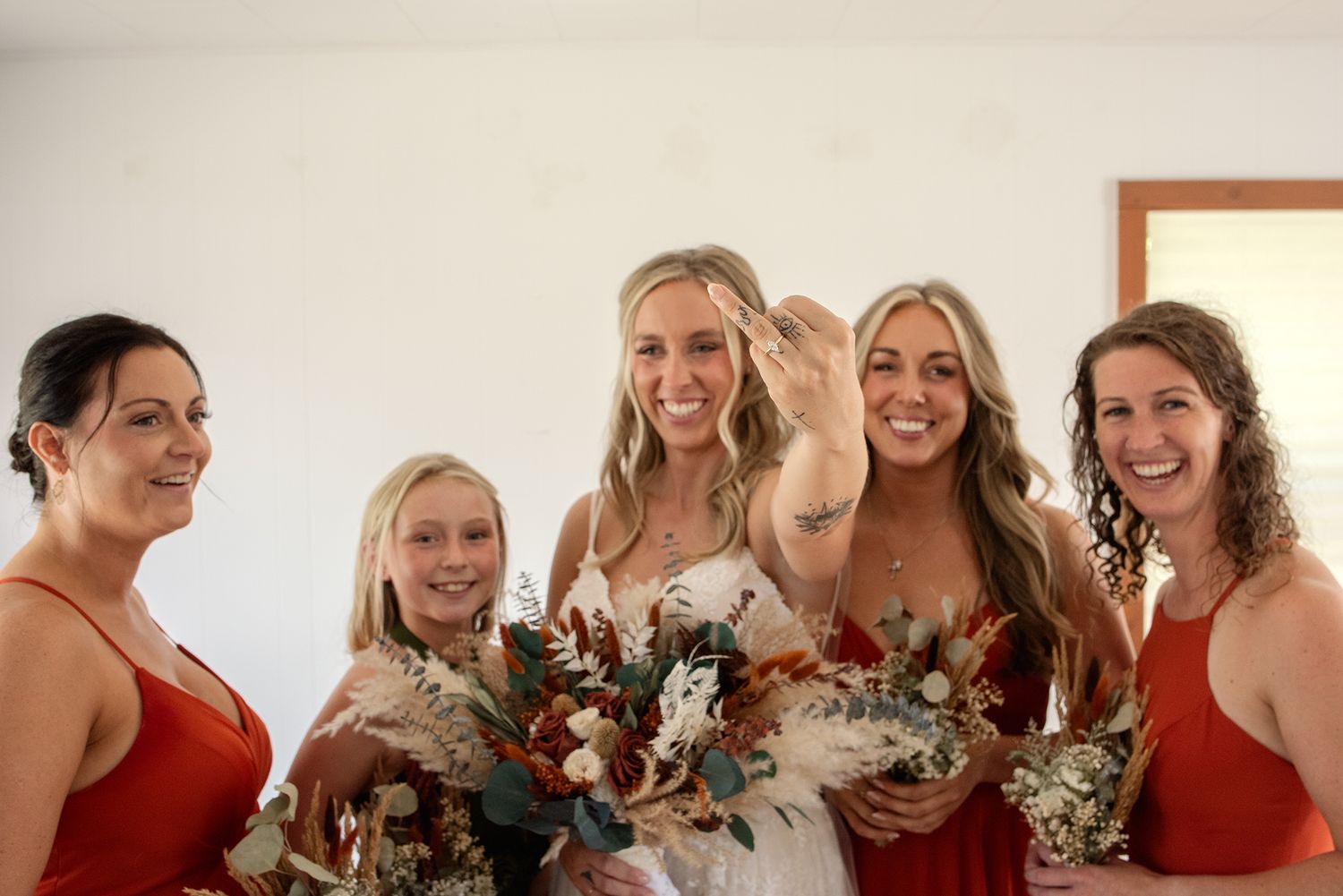 Bride showing off her ring with four bridesmaids in red dresses holding boho floral bouquets.