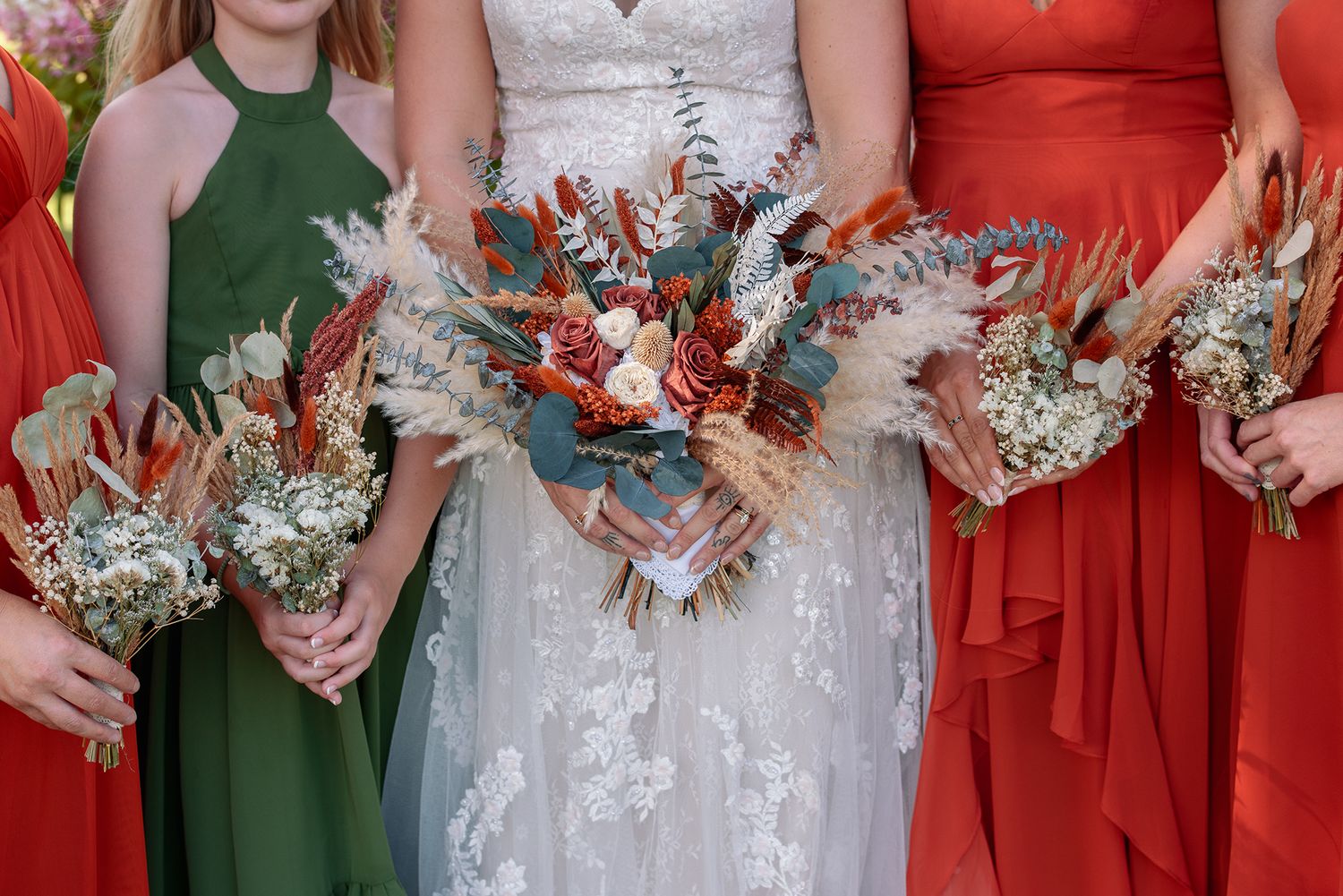 Bride and bridesmaids in green and red dresses holding rustic dried flower bouquets with eucalyptus and pampas grass.