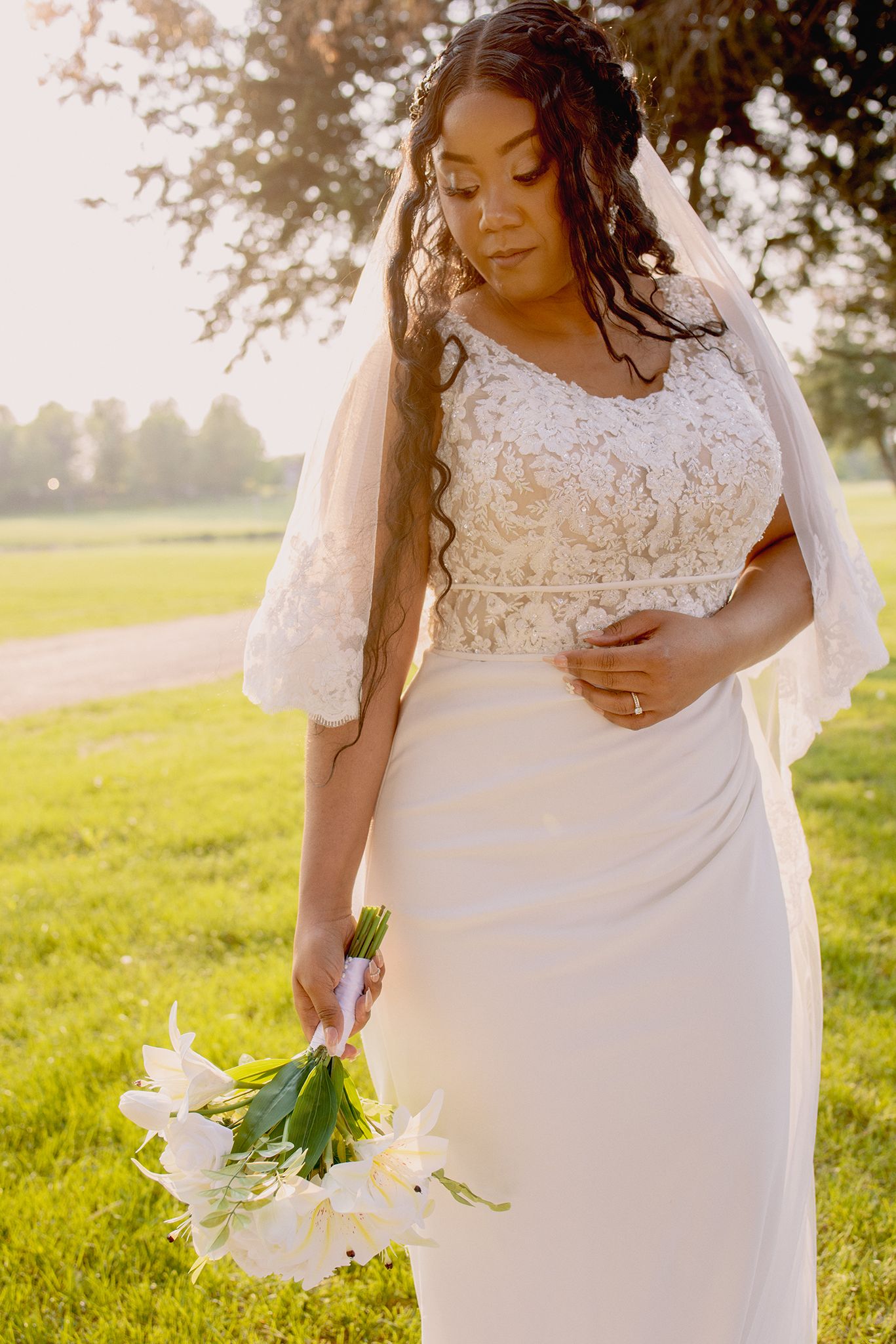 Bride in lace wedding gown holding white lily bouquet standing in sunlit park with veil flowing.