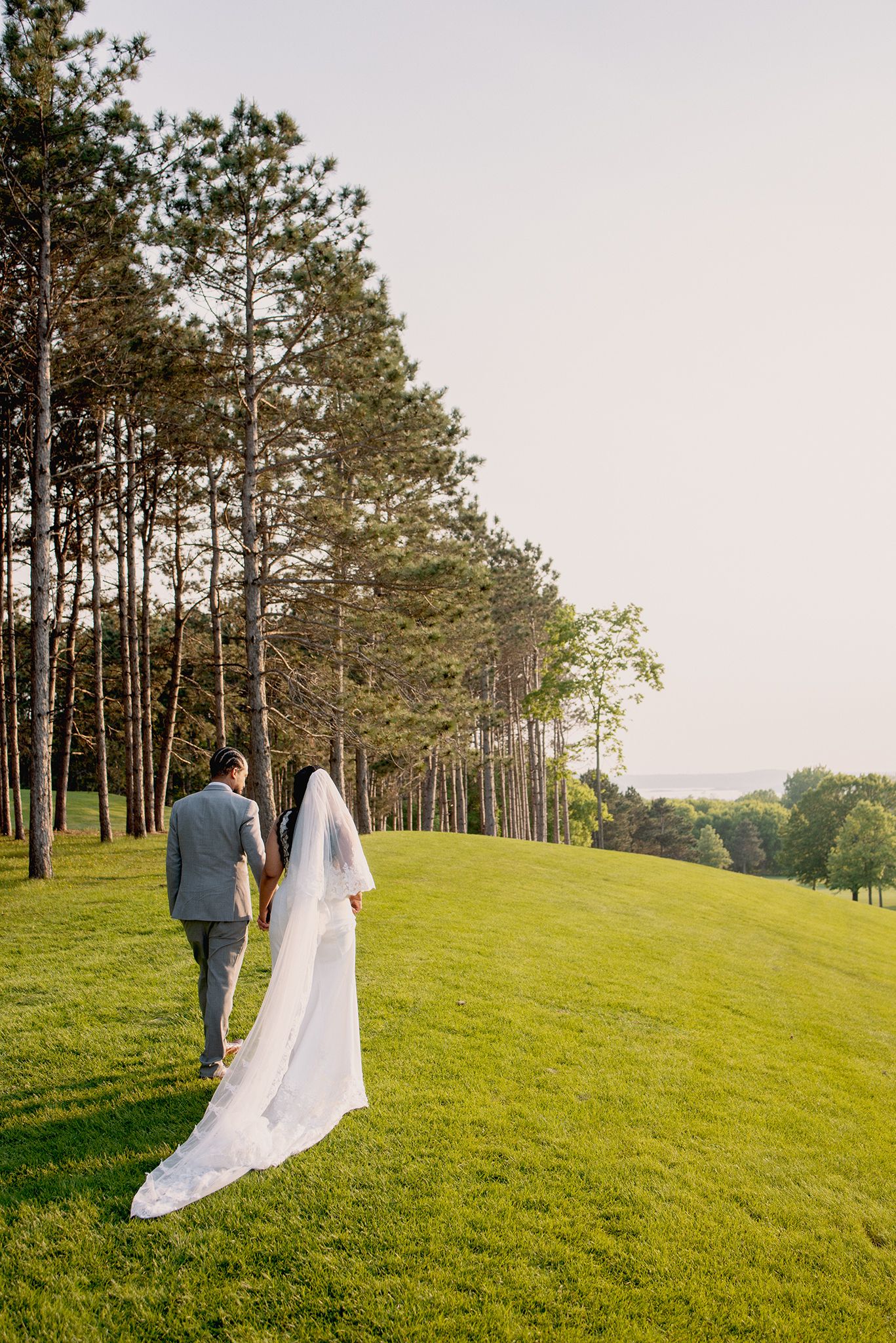 Bride and groom walking hand in hand across lush green golf course with tall pine trees.