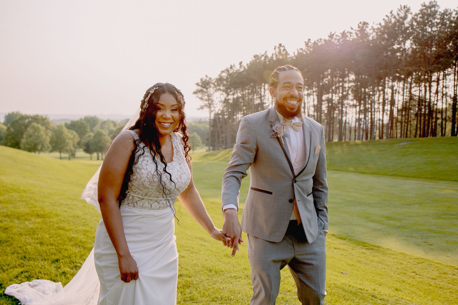 Happy couple holding hands walking through green field at golden hour after wedding ceremony.