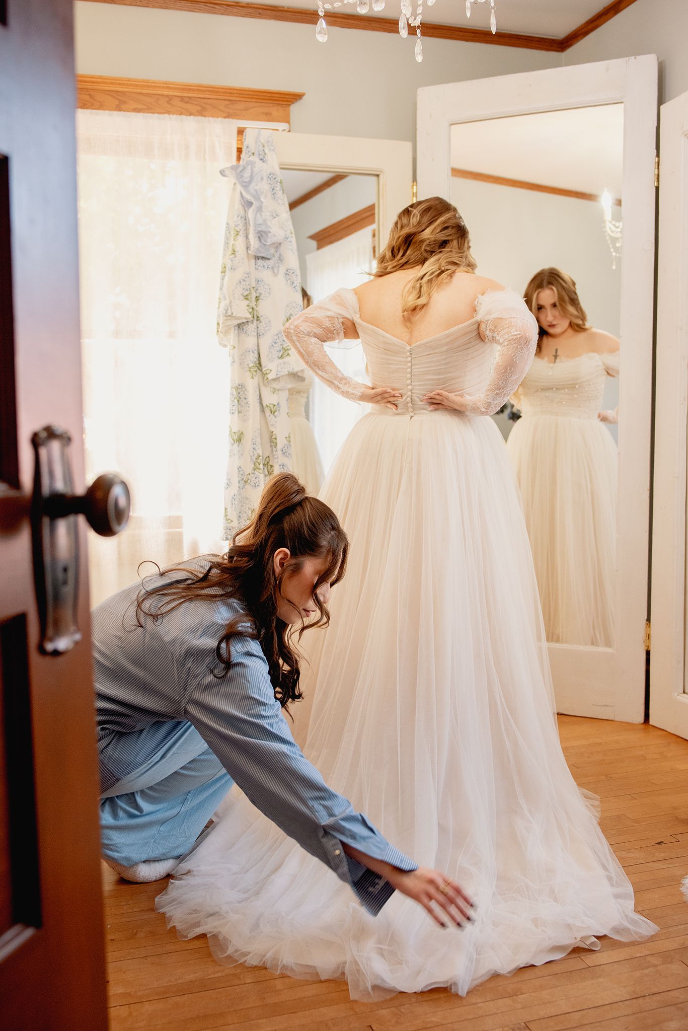 Assistant helping bride adjust white tulle wedding dress hem in bright dressing room with mirror.