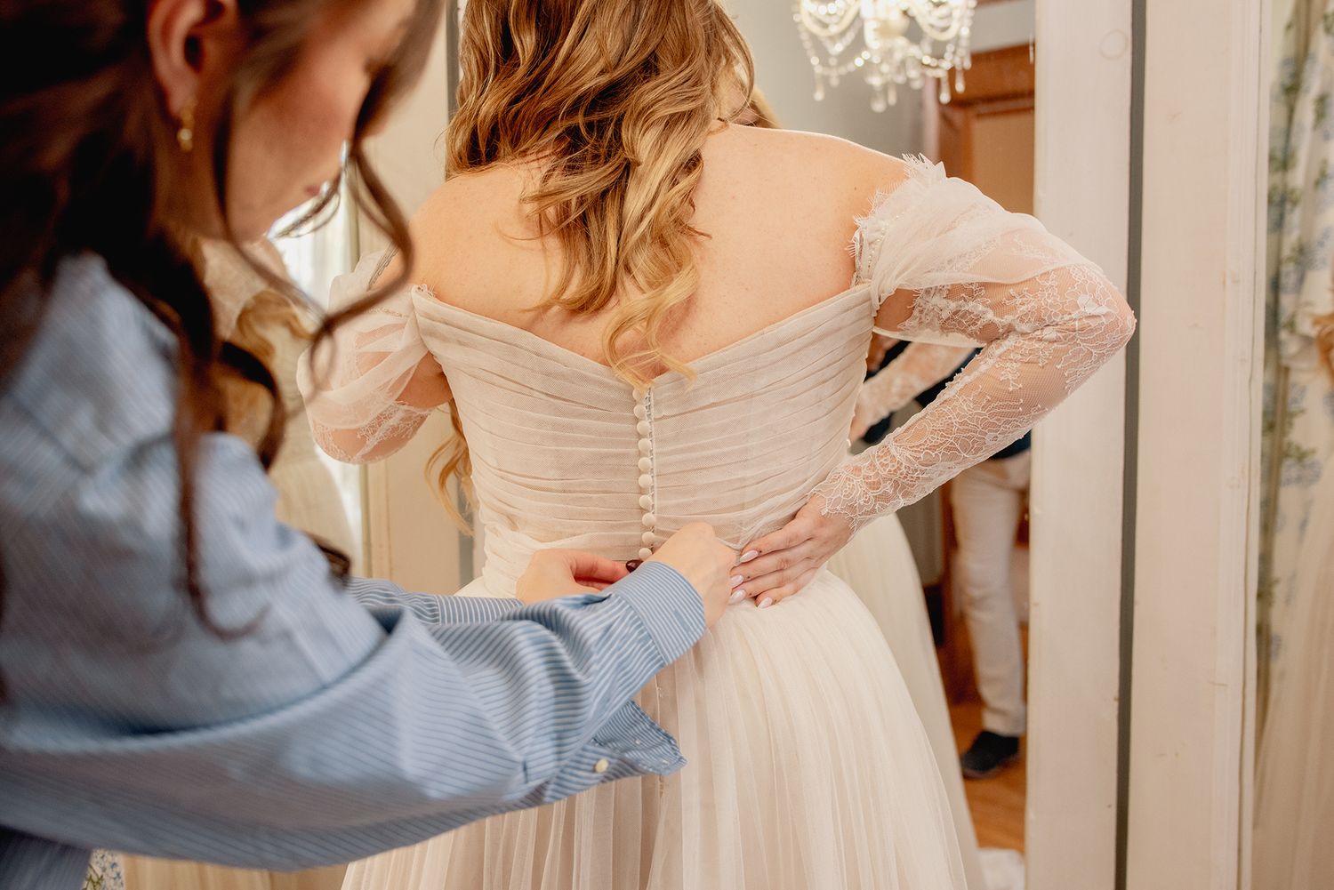 Bridesmaid helping bride button up elegant off-shoulder lace wedding gown from behind in bridal suite.