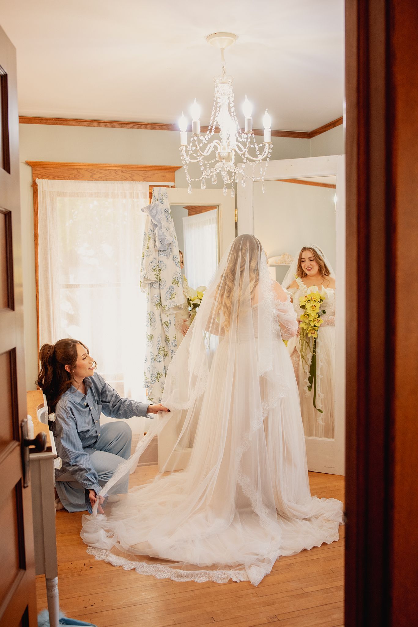 Bride in white gown and veil being helped by bridesmaid in blue, reflected in mirror with chandelier above.