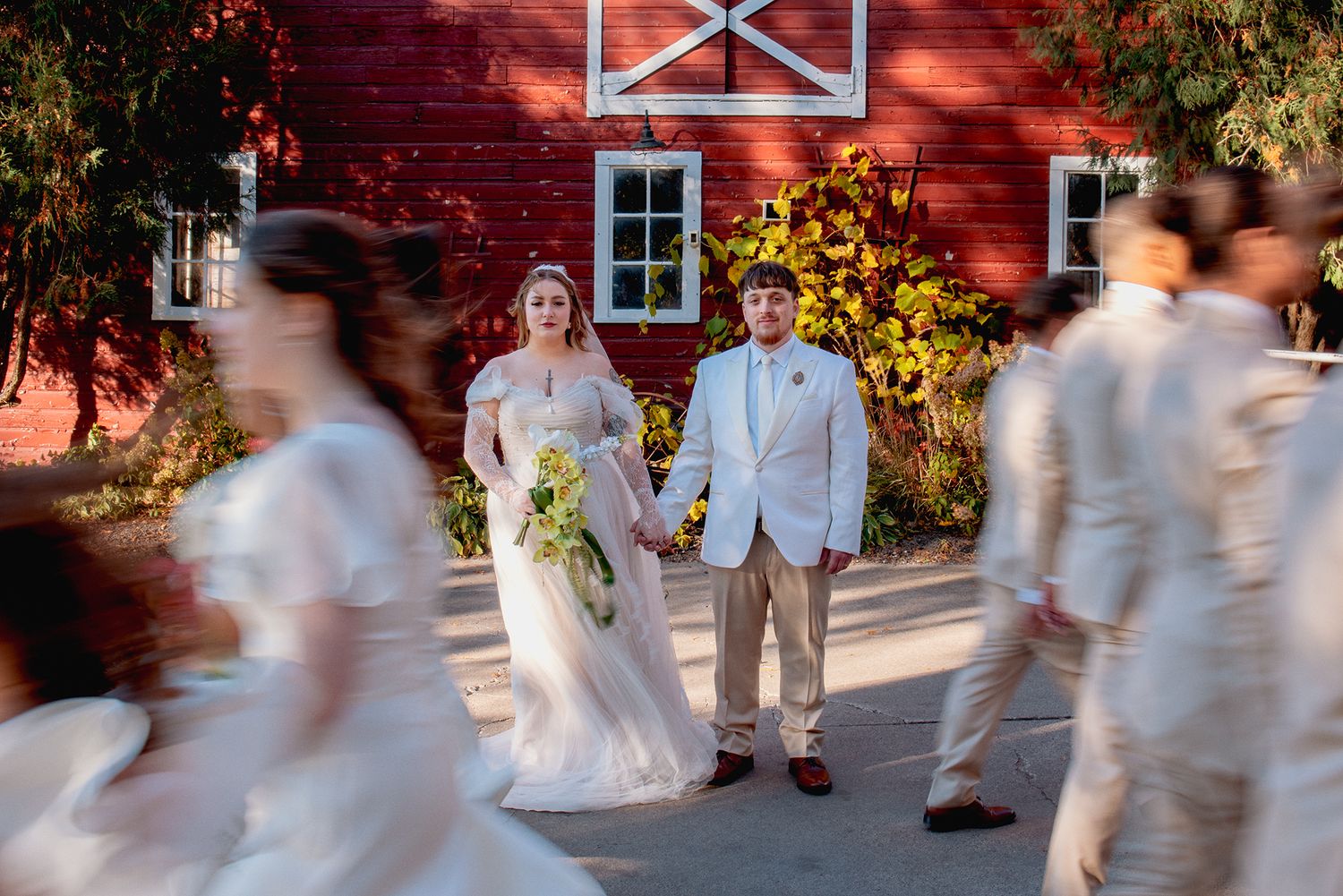 Bride and groom hold hands in front of red barn as wedding party walks past in blur.