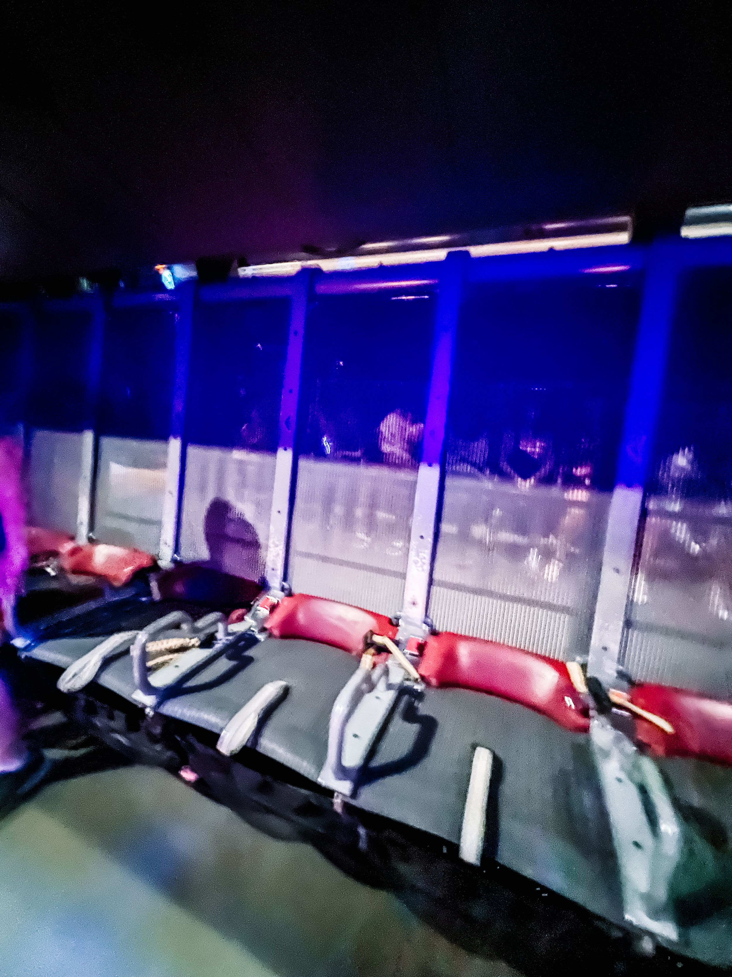 Hockey locker room benches with red padding illuminated by blue lighting and overhead lockers in dark environment.
