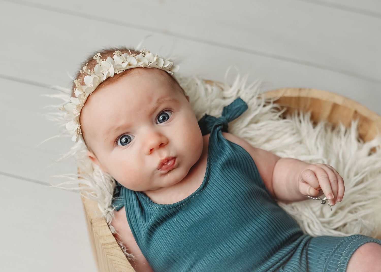 Baby wearing teal knit outfit lying on white textured blanket in professional photo shoot.
