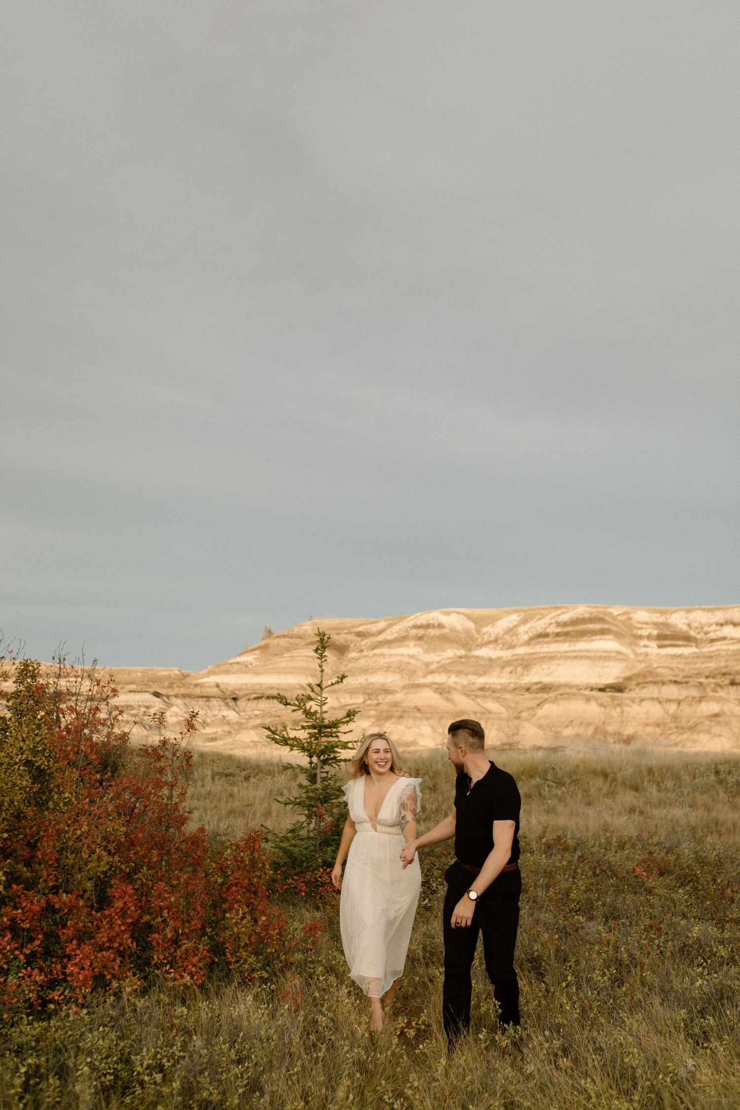 Couple walking through prairie field with dramatic rocky cliffs in background at sunset.