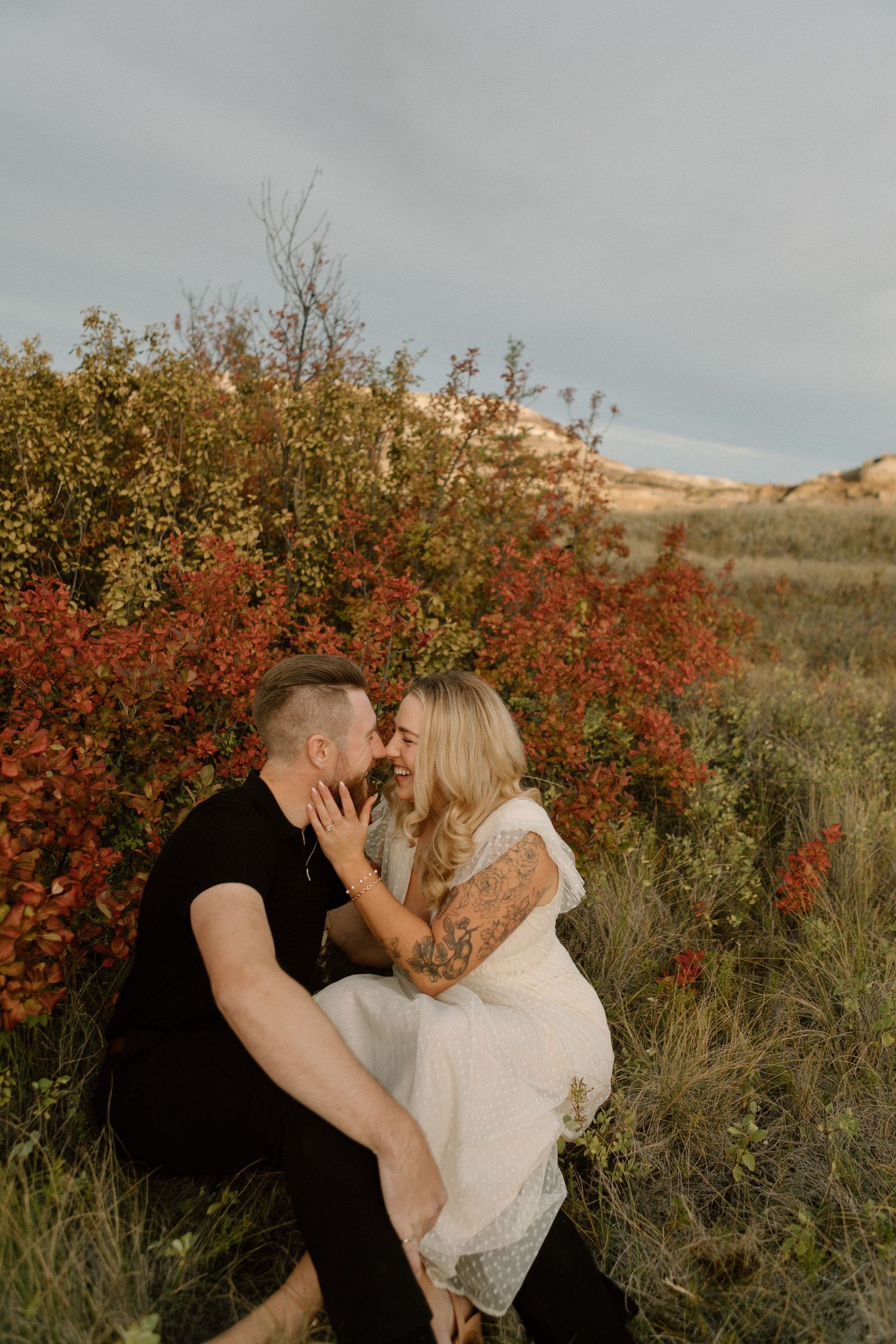 A couple sits together intimately in a field with autumn red bushes and golden hills in the background.